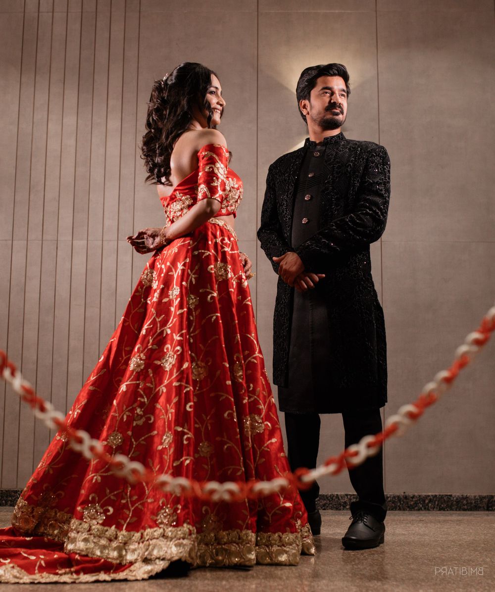 Elegant couple in red and black formal Indian wedding attire posing against neutral backdrop.