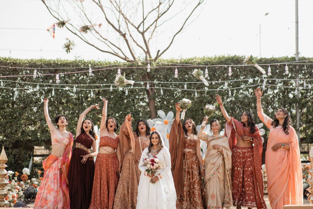 Wedding party in peach and rust colored dresses celebrating with raised arms against garden backdrop.