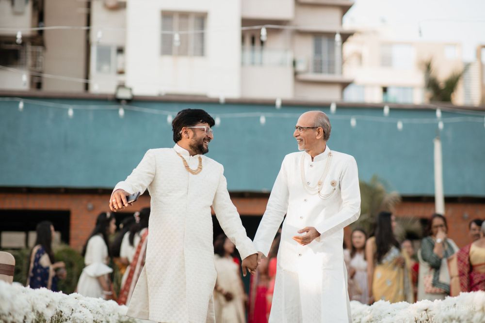 Two people in matching white traditional outfits hold hands during an outdoor celebration.