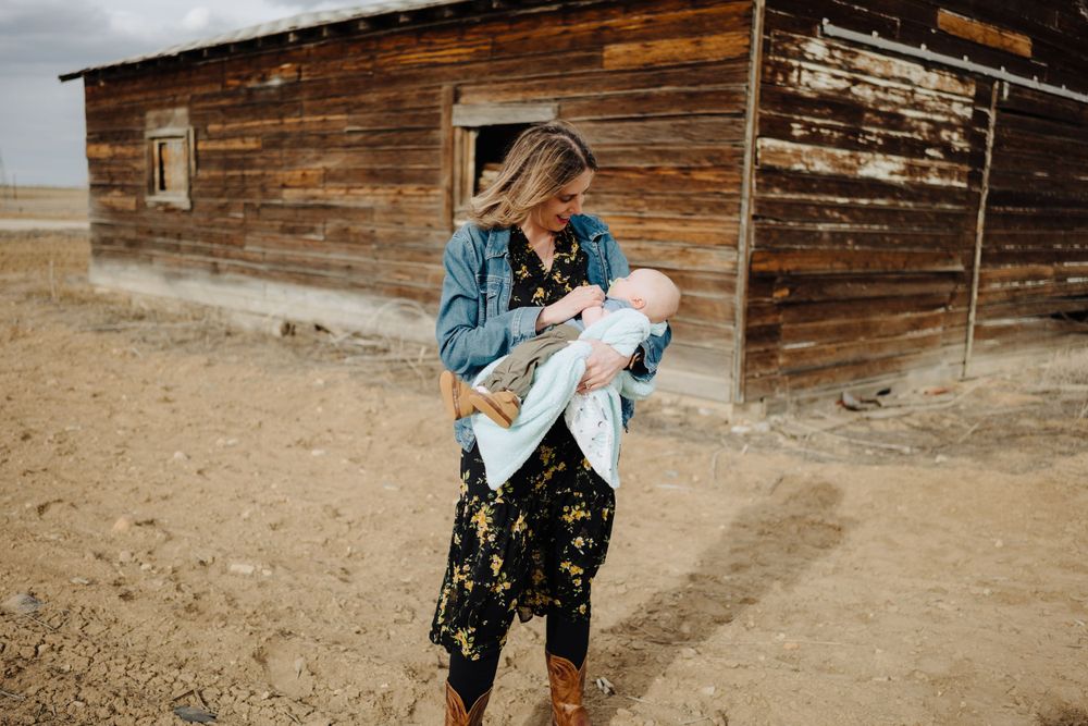 Woman in denim jacket holds infant outside weathered wooden barn in rural setting.