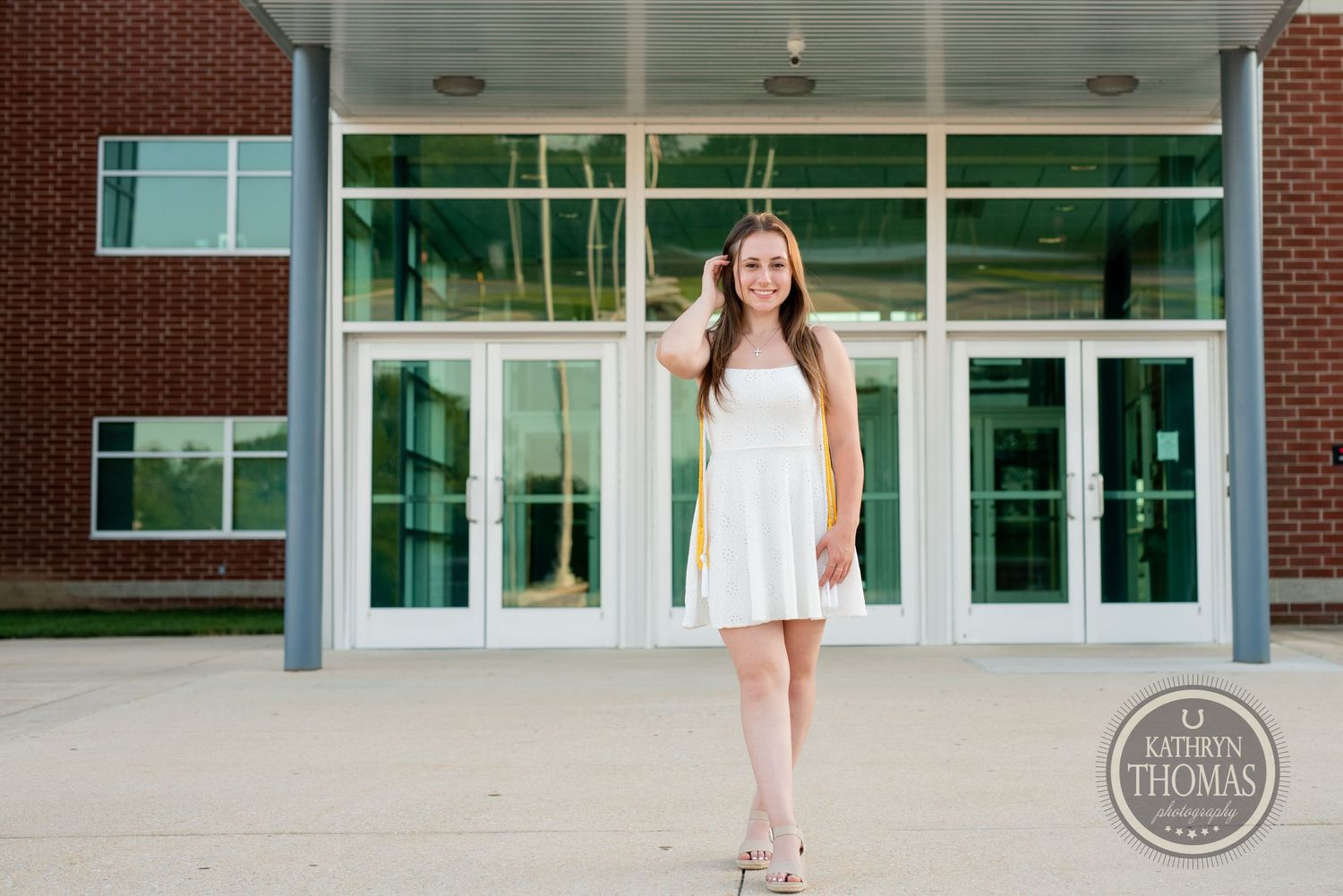 Graduate in white dress stands in front of modern brick building with glass doors.
