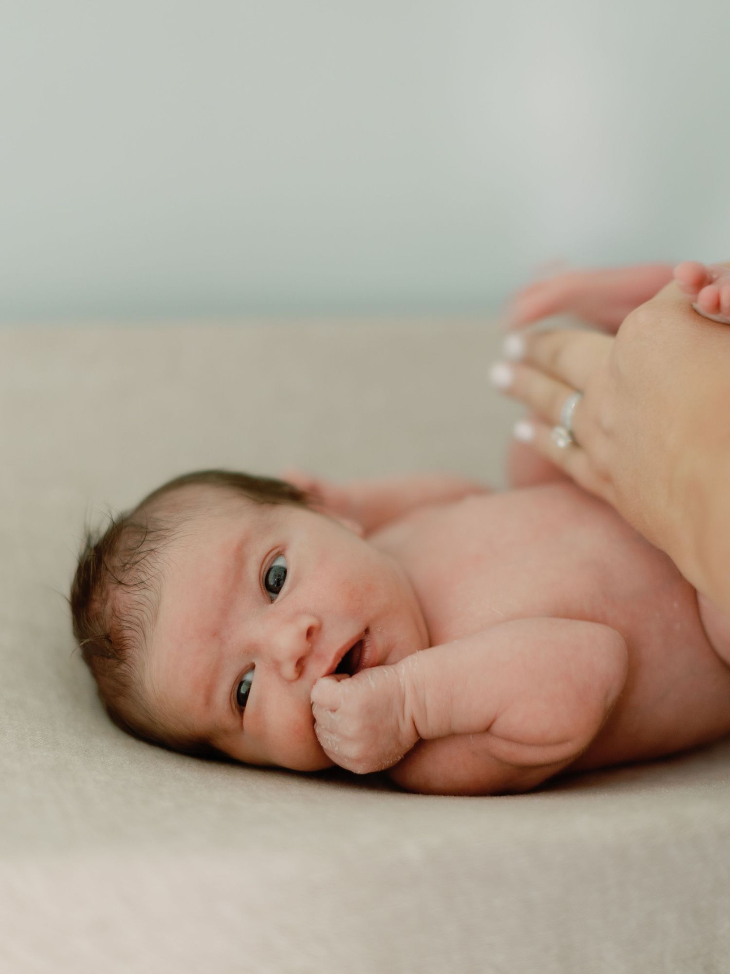 Newborn baby lying on soft beige surface sucking thumb with gentle smile.