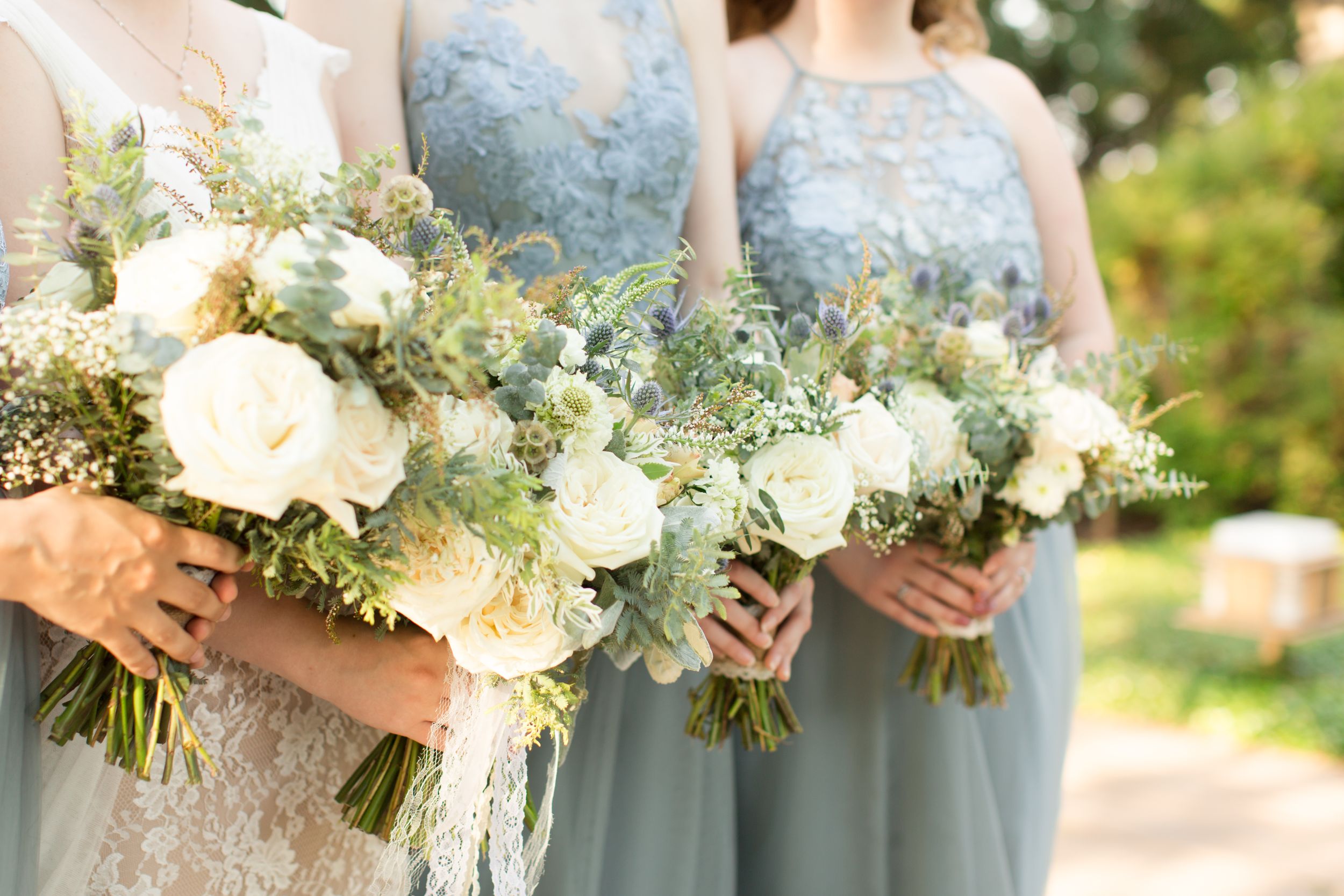 Bridesmaids in light blue dresses hold white and green floral bouquets outdoors.