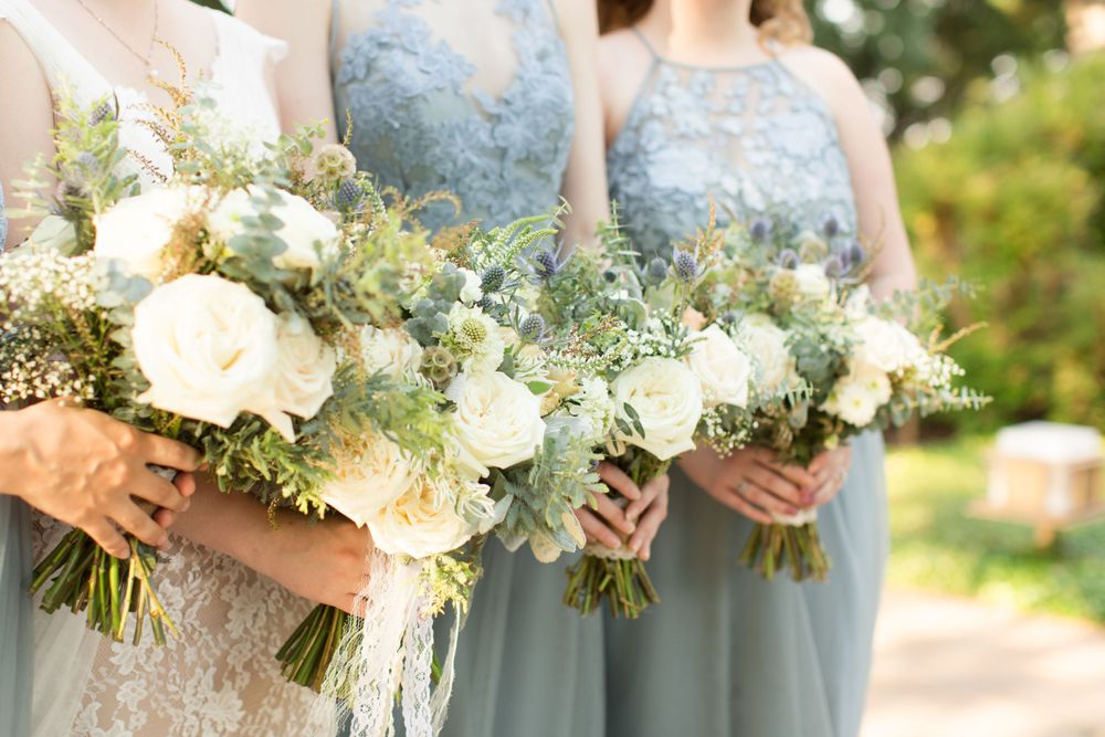 Bridesmaids in light blue dresses hold white and green floral bouquets outdoors.