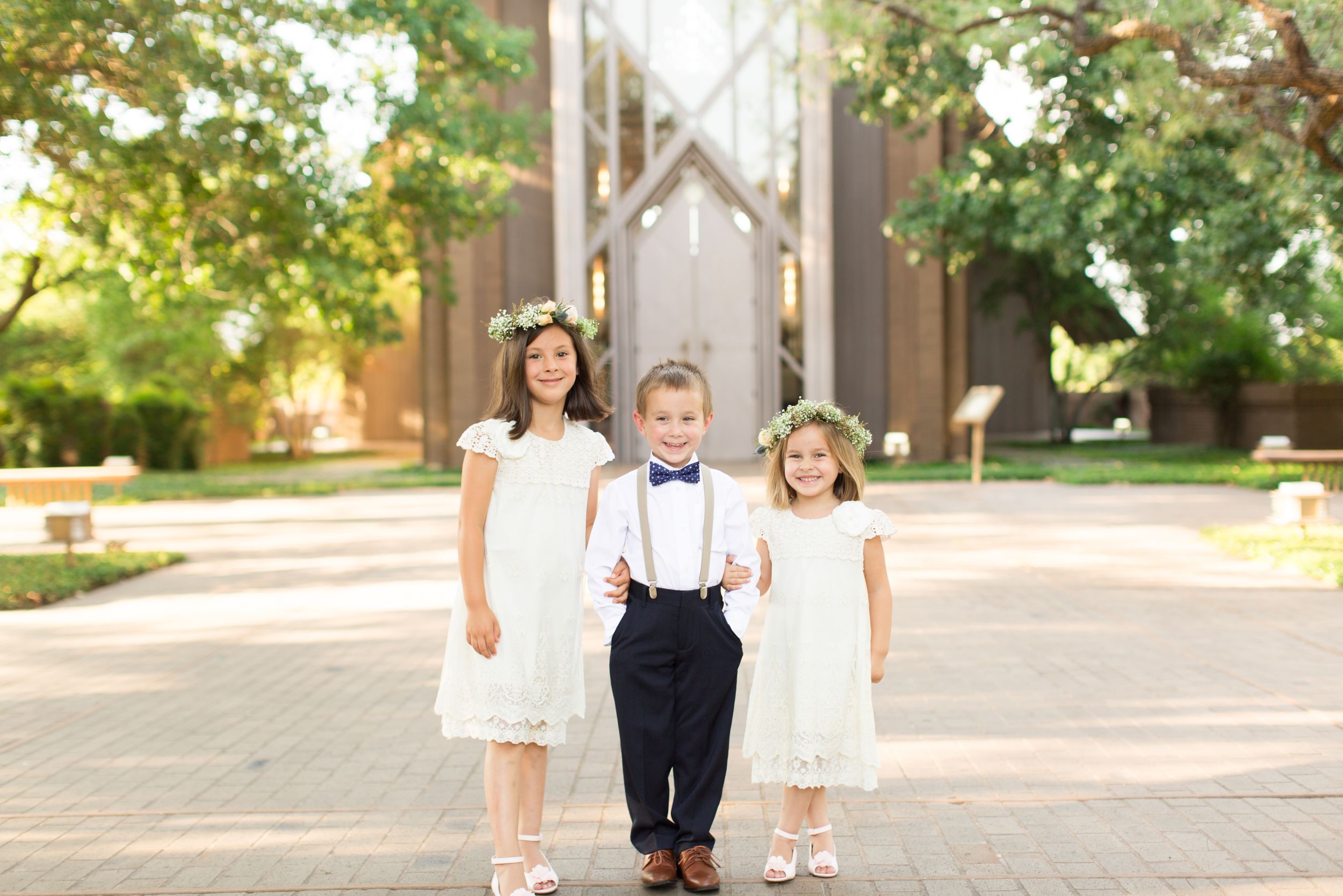 Ring bearer and flower girls walk hand in hand on a sunlit outdoor path wearing formal attire.