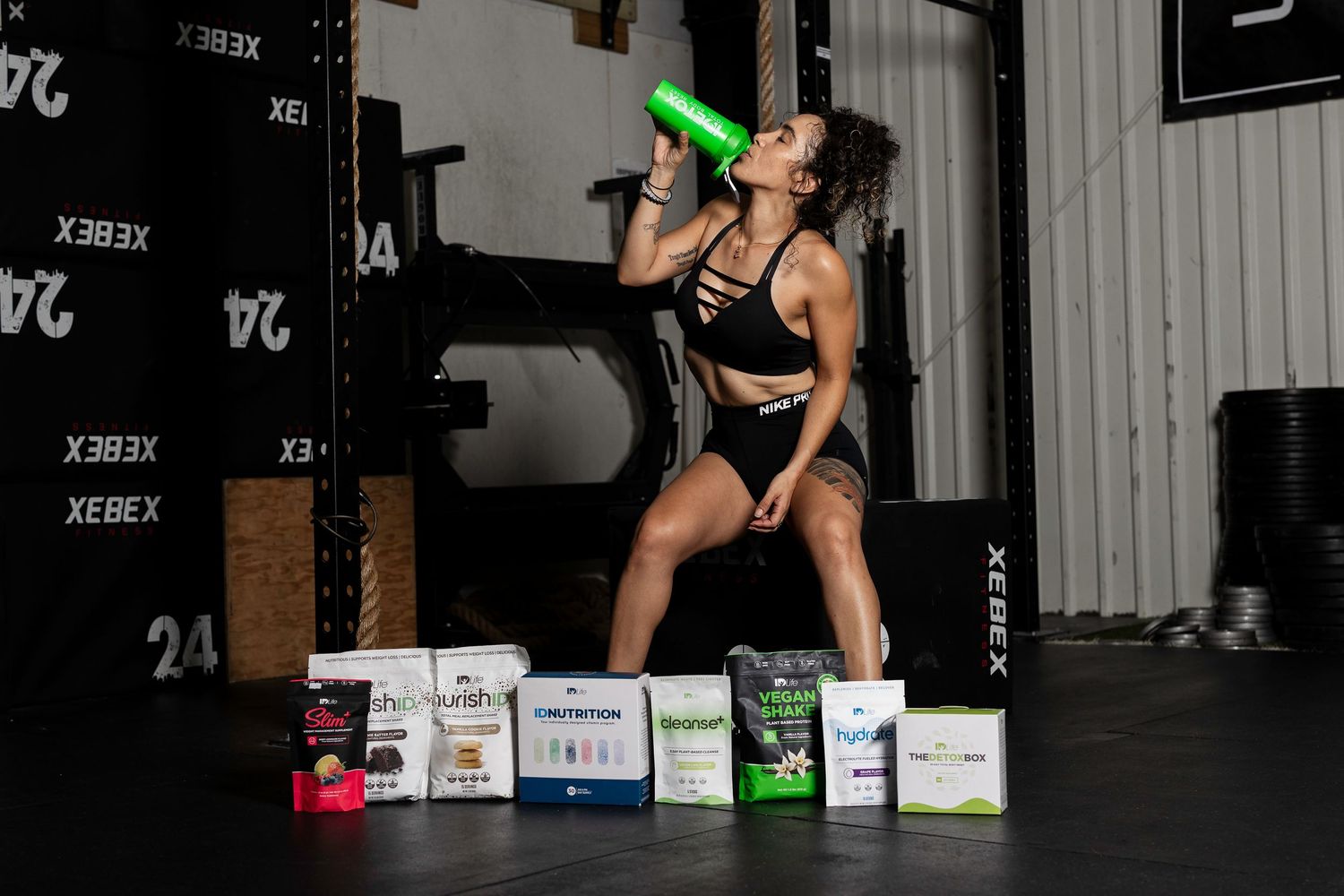 Fitness enthusiast drinking from bottle surrounded by supplement products in gym setting.