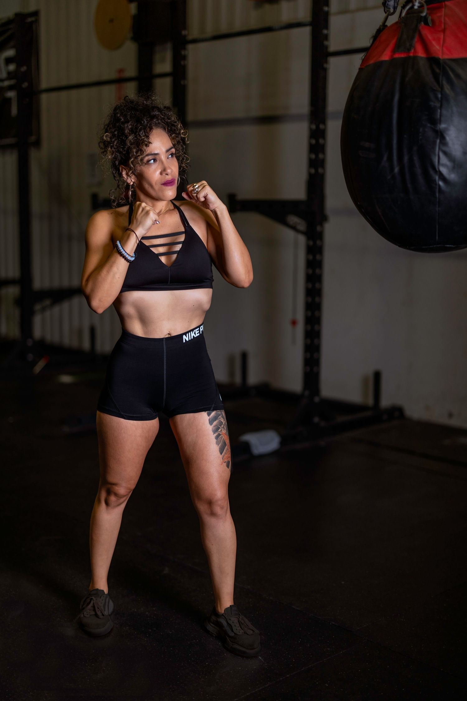 Athletic woman in workout attire strikes boxing pose next to punching bag in dimly lit gym.