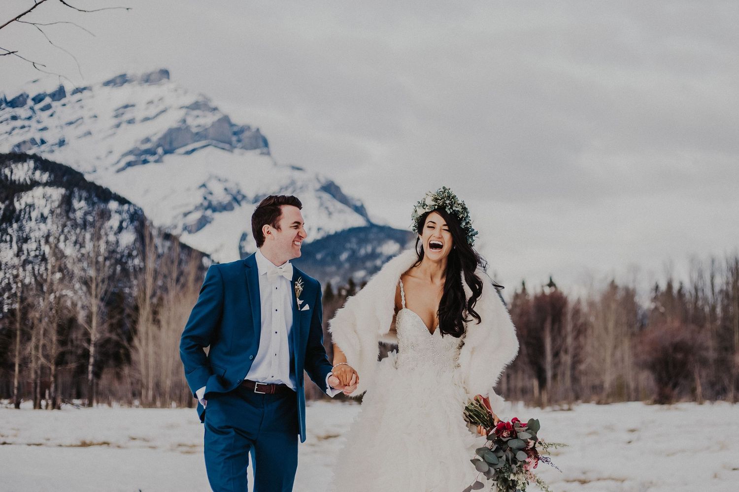 Bride and groom holding hands and laughing walk in front of the Banff mountains