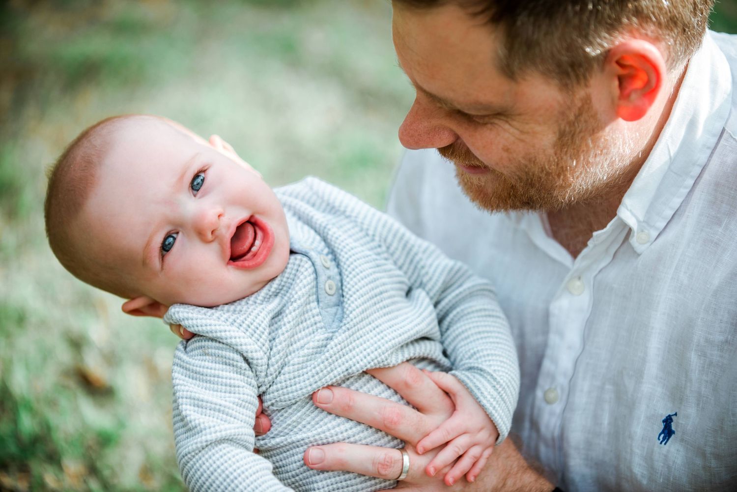 Joyful interaction between adult and smiling baby wearing a light blue sweater outdoors.