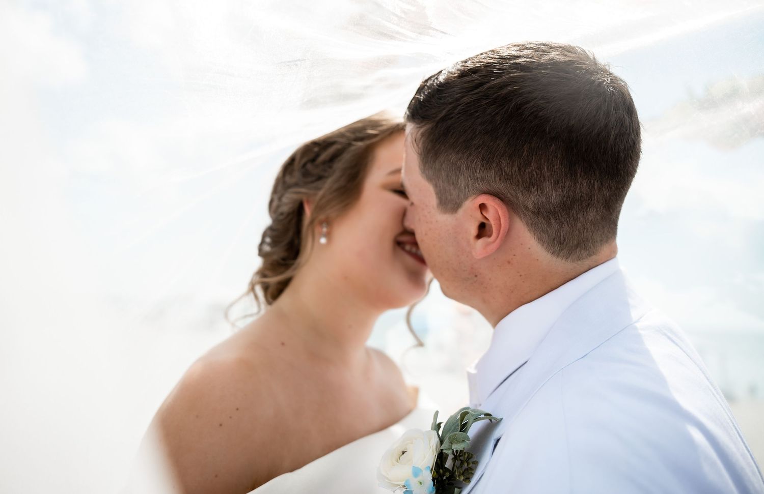 Bride and groom sharing a romantic kiss under a flowing white veil outdoors.