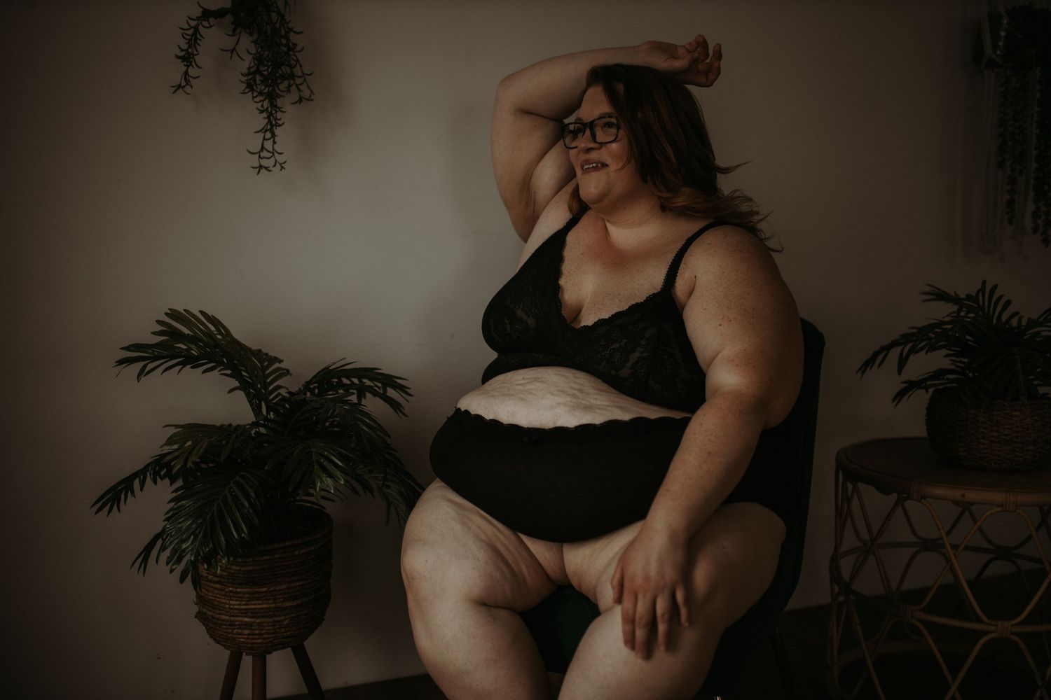 A body-positive boudoir portrait in moody lighting with indoor plants and natural decor elements in the background.
