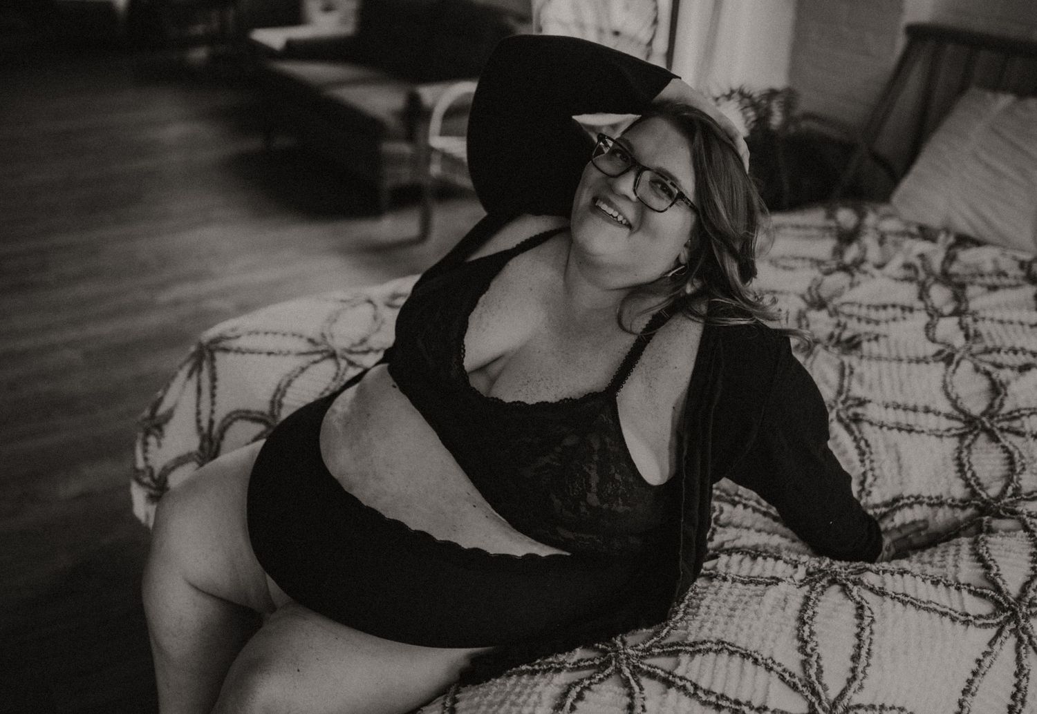 Black and white boudoir photo of a smiling plus sized woman in underwear posing on patterned bedding.