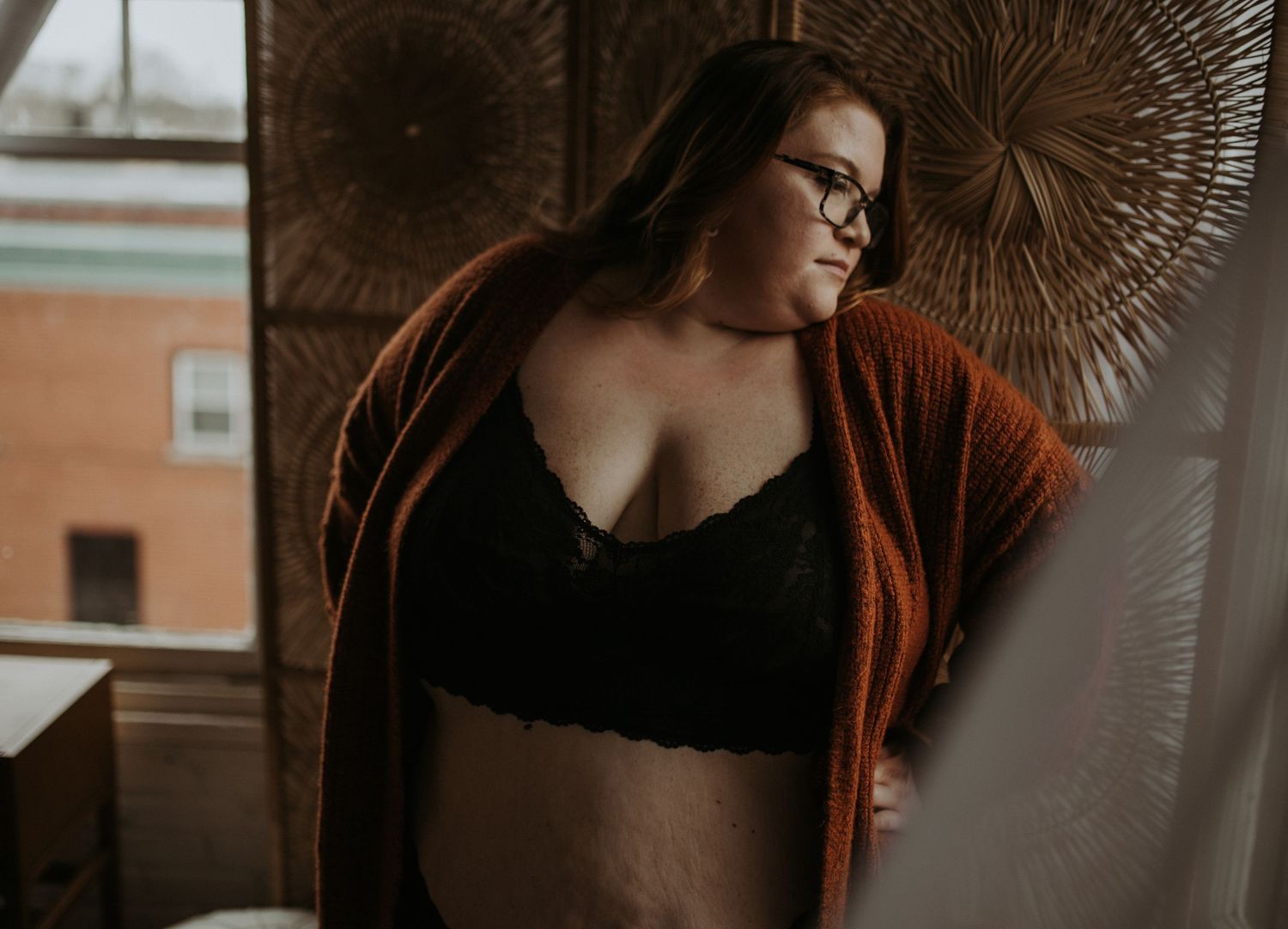 A woman wearing glasses, a rust-colored cardigan, and black lace top sits near window with rattan sunburst decor in background.