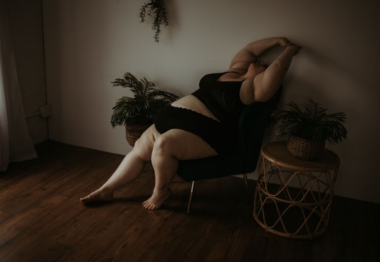 A moody boudoir photo of a curvy woman posing creatively on wicker furniture and houseplants in a dimly lit room.