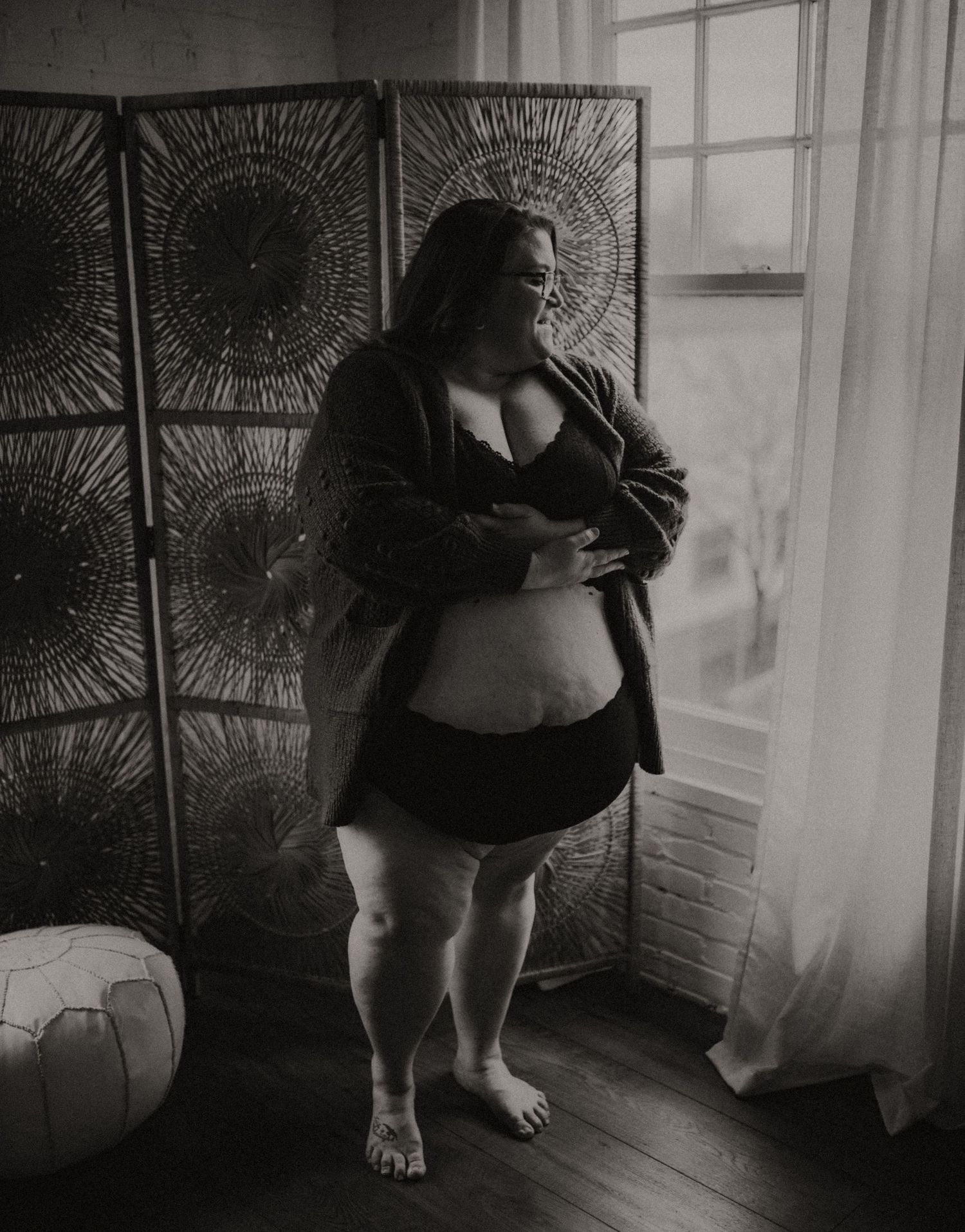 Black and white artistic boudoir photograph showing a plus size woman in casual lingerie near a window with decorative screens.