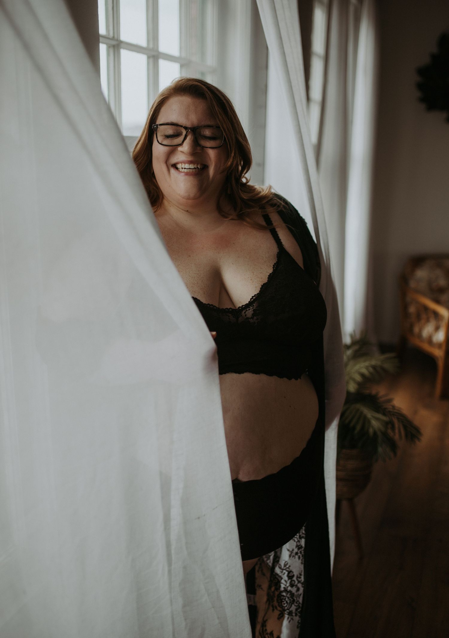 A woman posing joyful while wearing glasses and black lingerie poses between white curtains by a window.