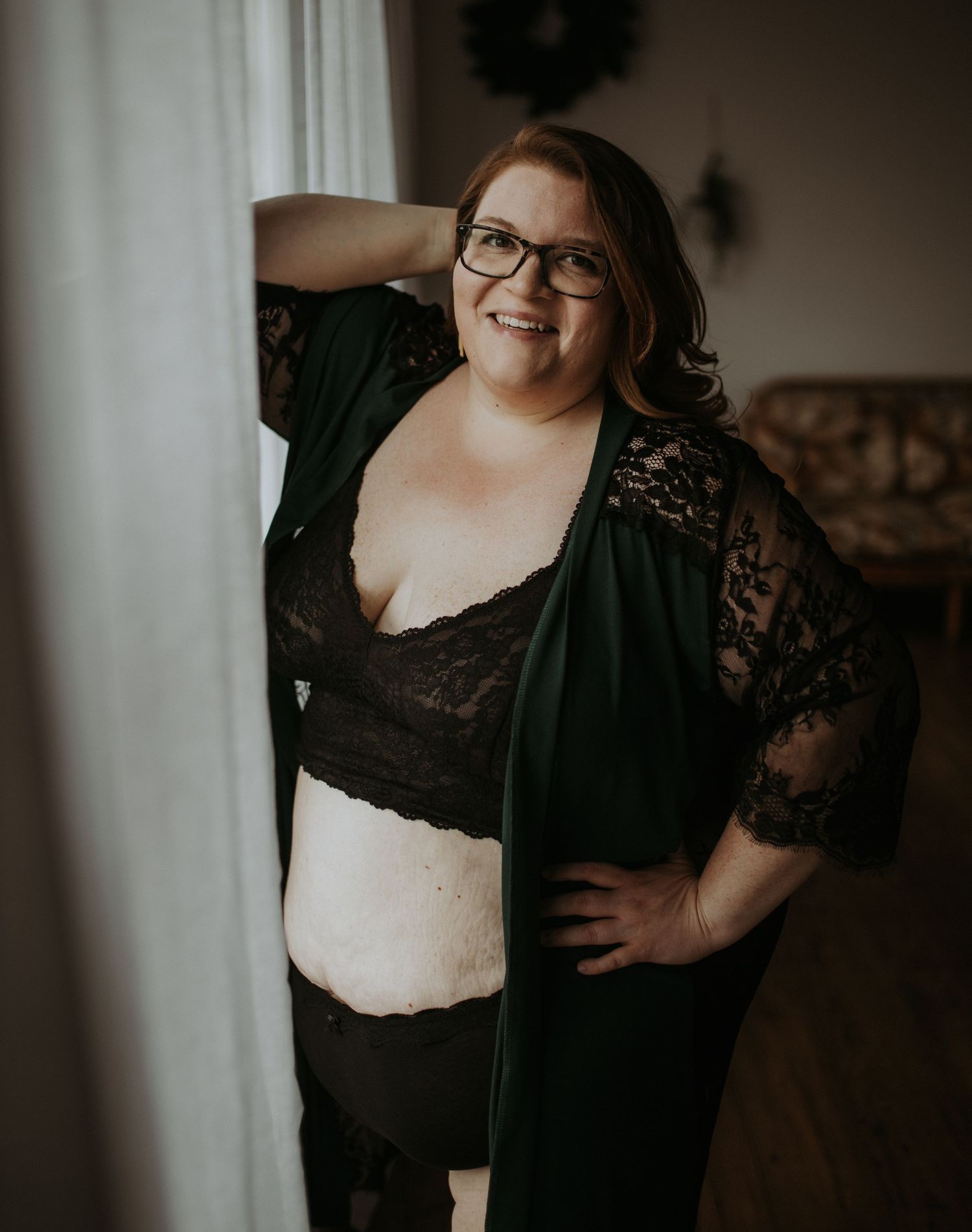 A plus size woman wearing glasses poses confidently in black lace lingerie and a dark cardigan against an indoor backdrop.