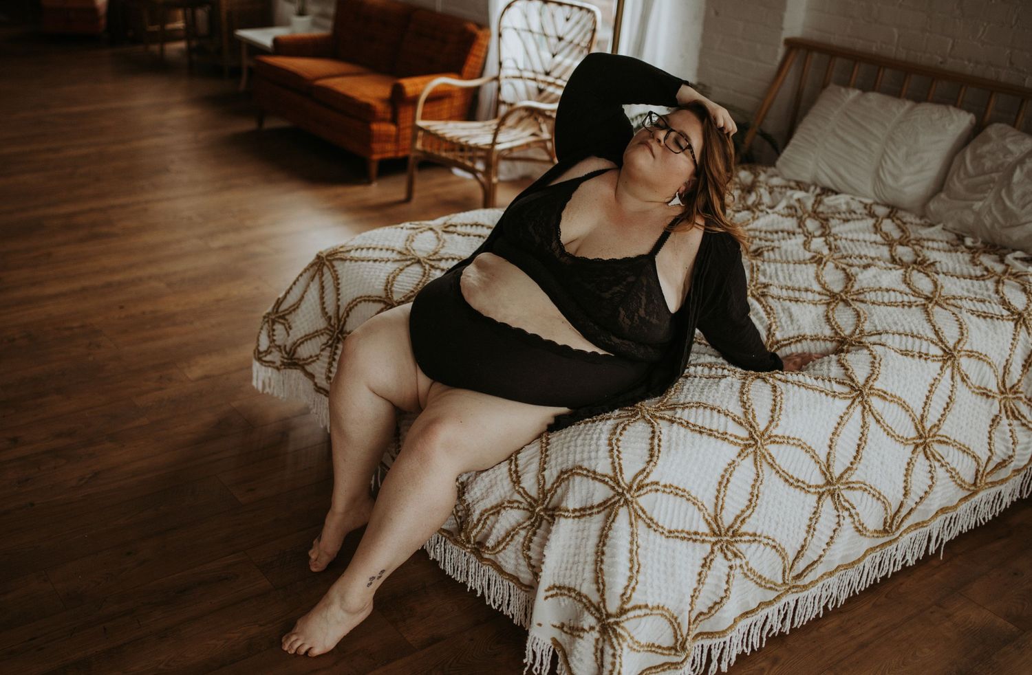 A plus sized woman in black loungewear reclines on a bed with hardwood flooring visible in the warm-toned room.