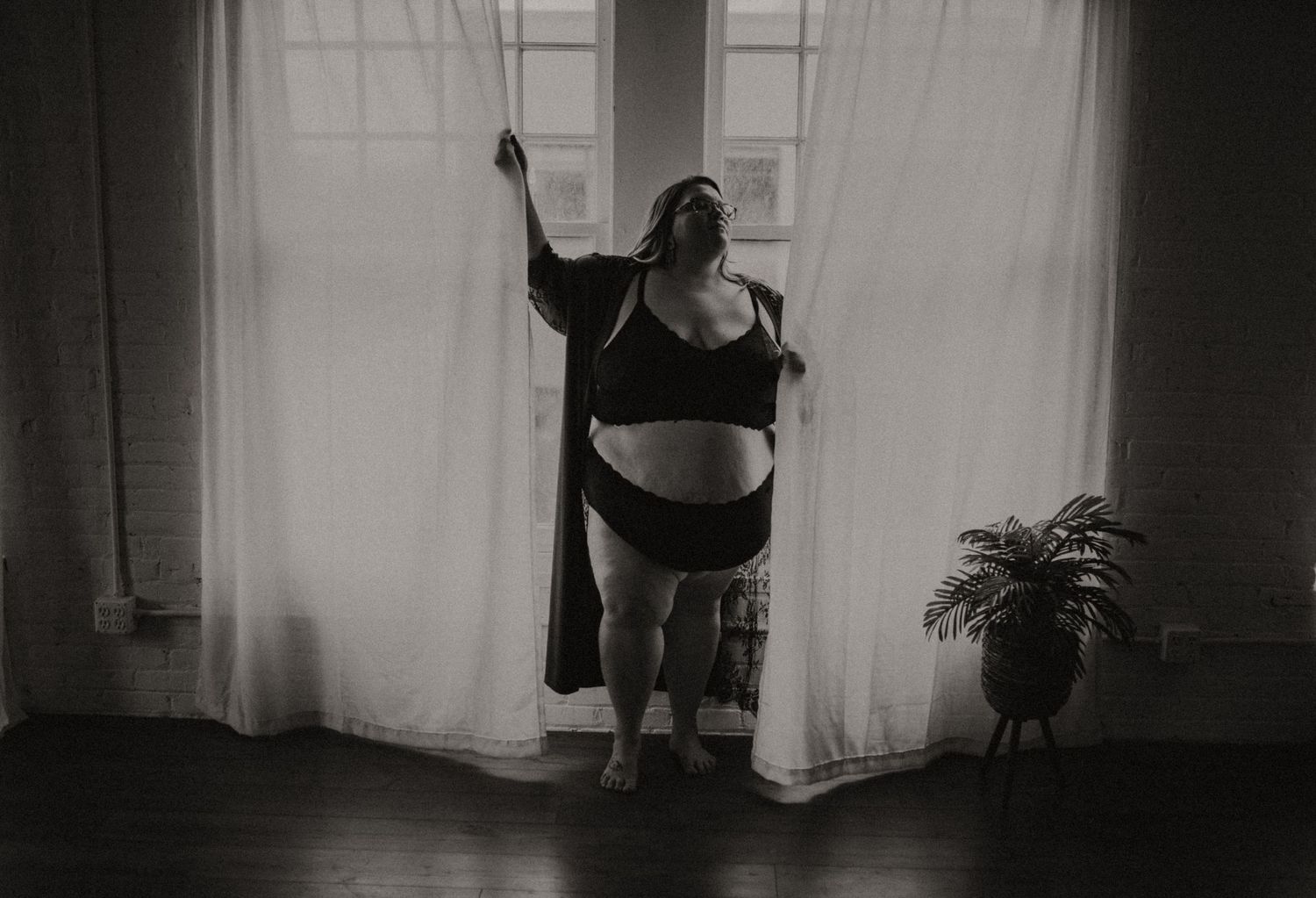 A silhouette of a woman in black lingerie stands gracefully in front of sheer curtains with sunlight streaming through the window.