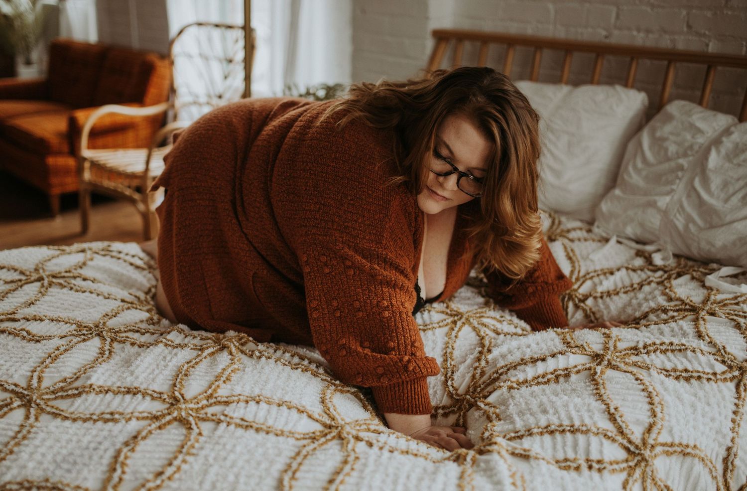 A plus sized in a cozy rust-colored sweater relaxes on a bed with a geometric patterned comforter in warm lighting.