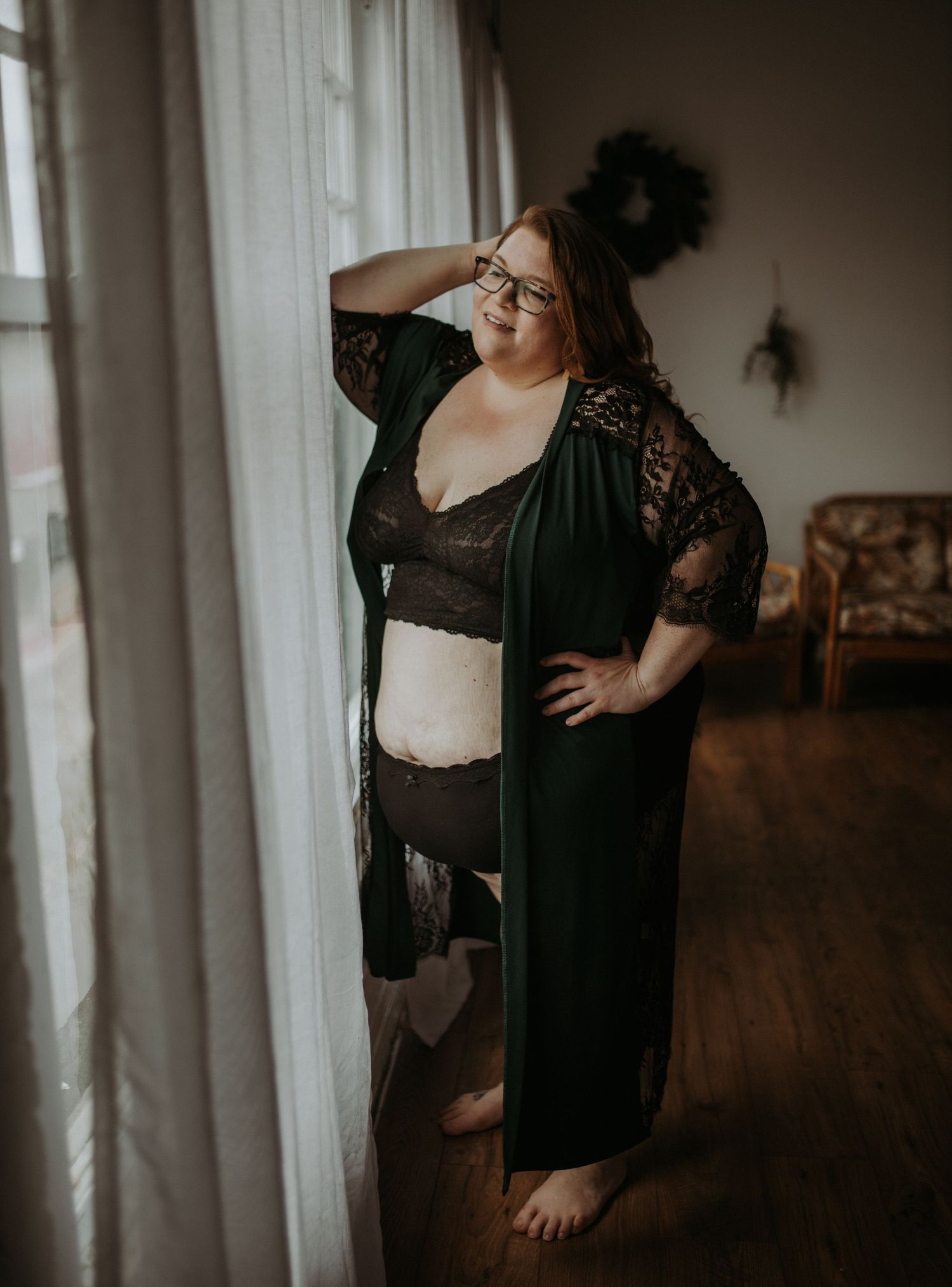 A boudoir photo shoot features a curvy woman in black lace lingerie and a sheer robe looking longingly by a window.