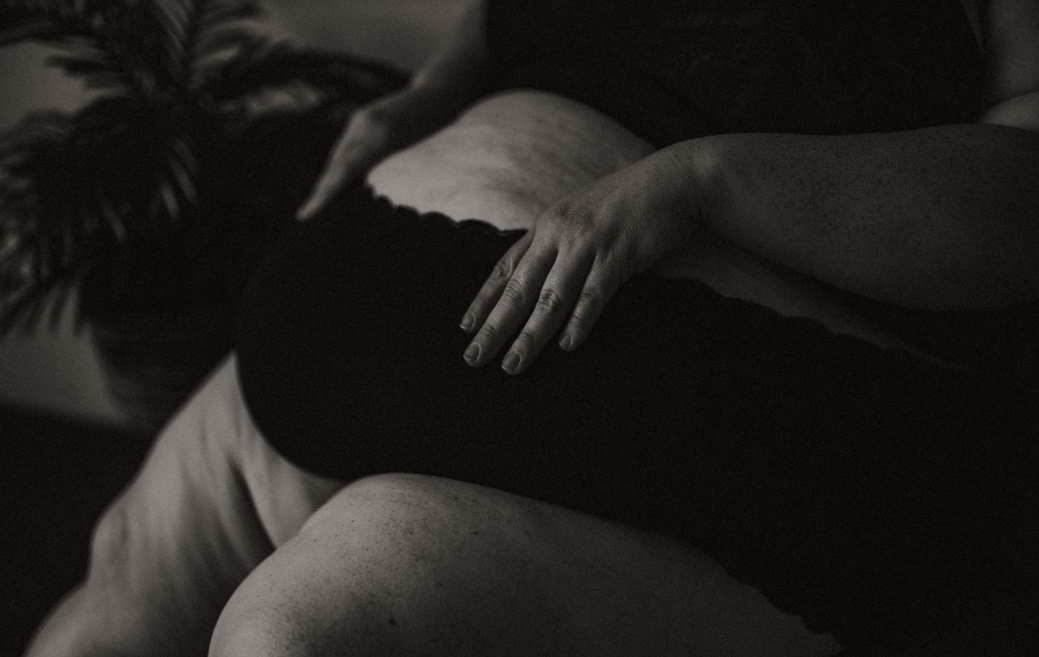 Black and white photograph of a plus size woman resting her hand on their belly in soft light.