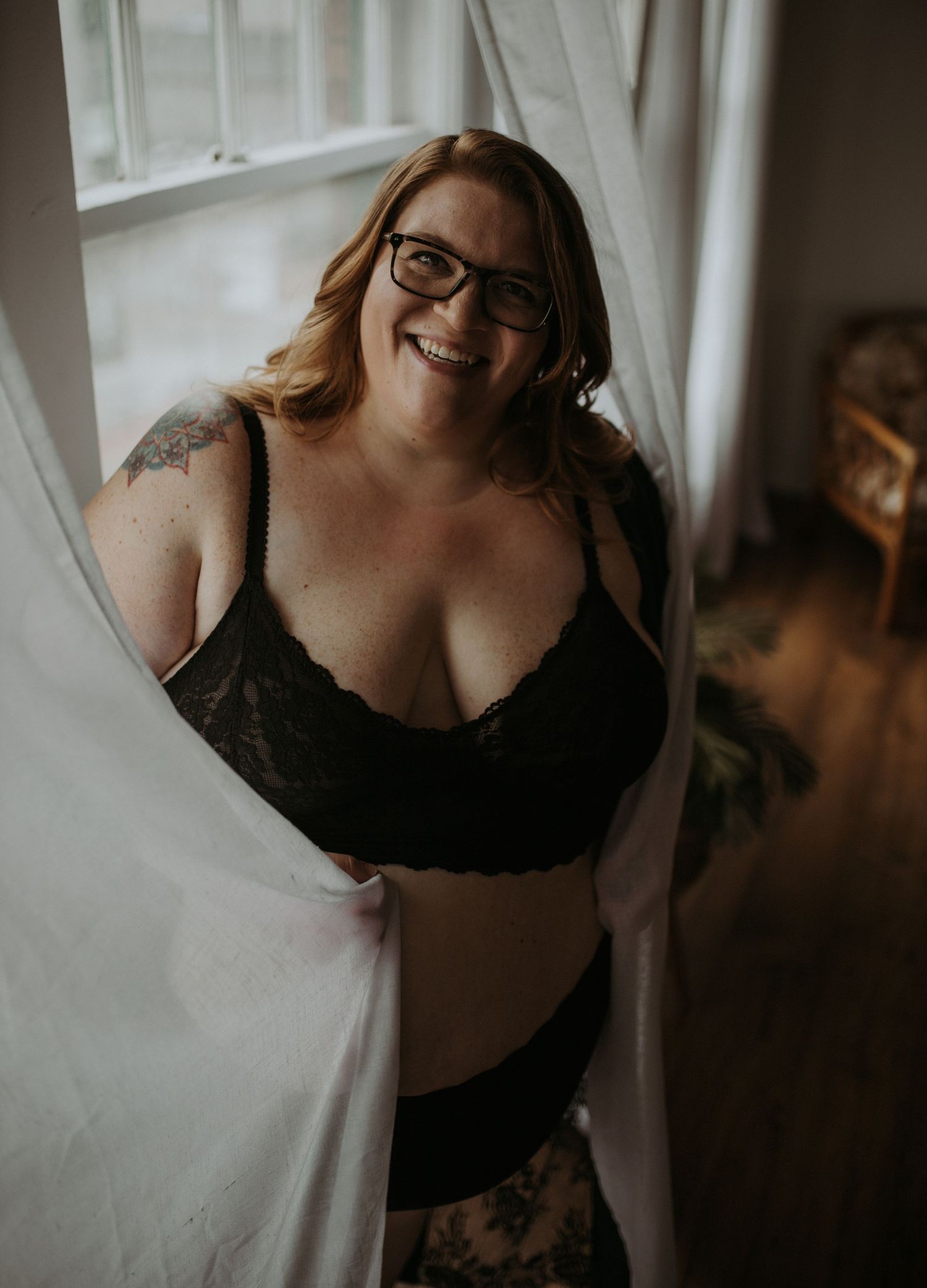 A tasteful boudoir portrait in monochrome features a plus size woman smiling in black lingerie posing near a window with sheer curtains.