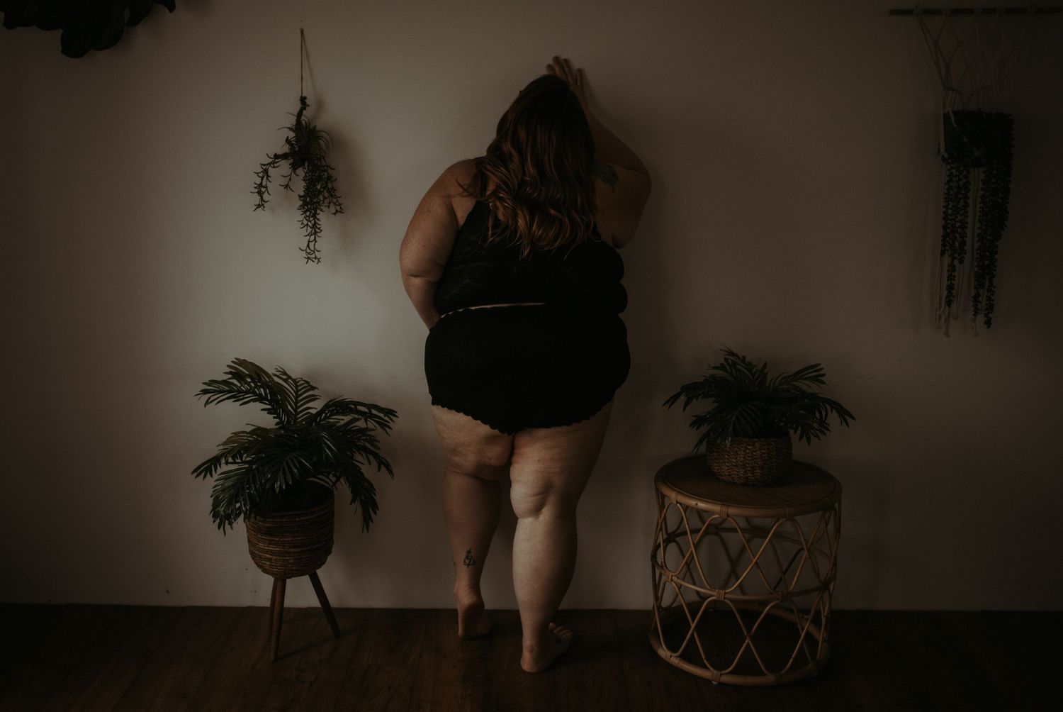 Artistic photo of a woman standing between two potted plants against a dark wall in moody indoor setting.