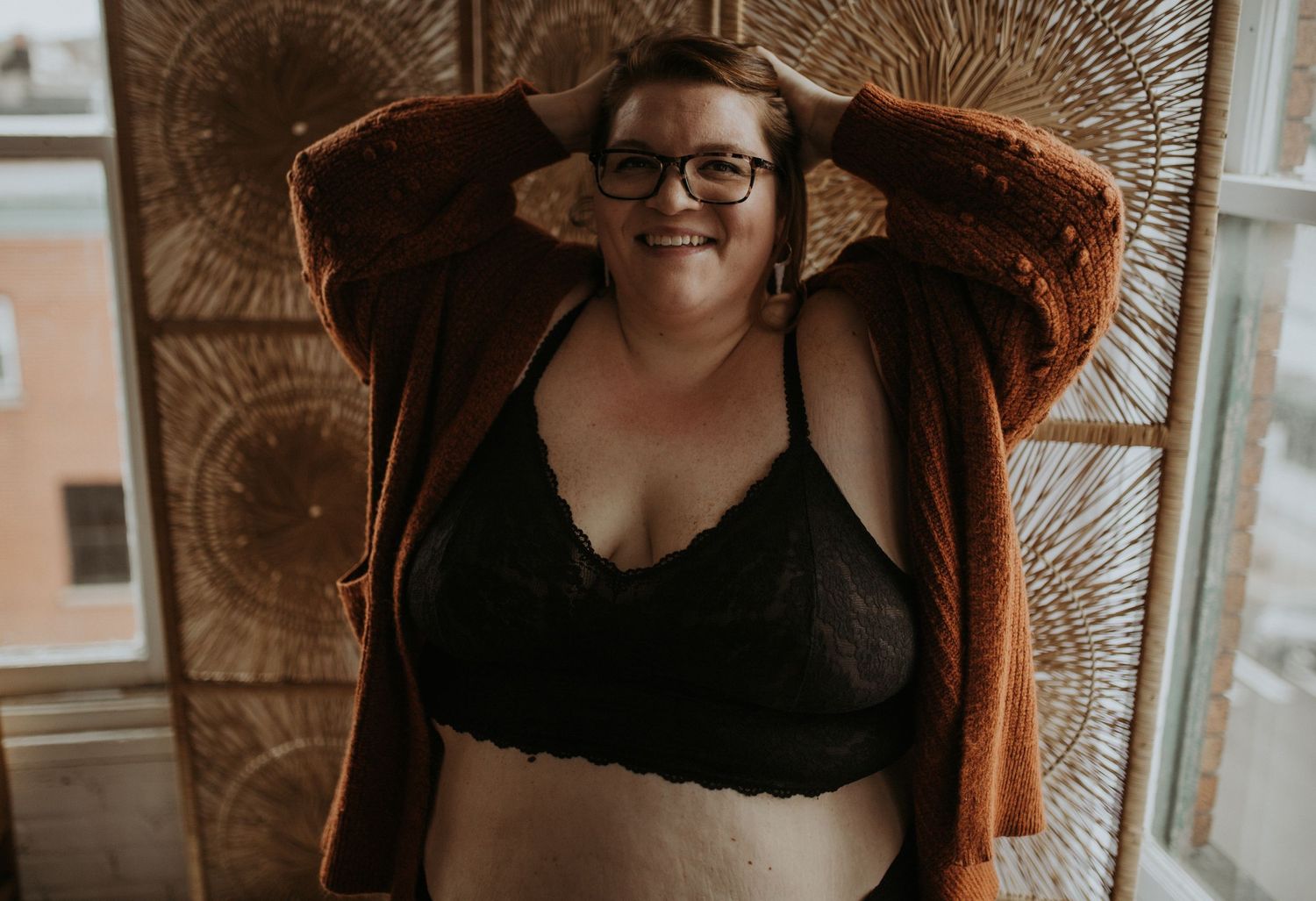 A woman wearing a rust-colored cardigan and black bralette poses confidently against a patterned window background.