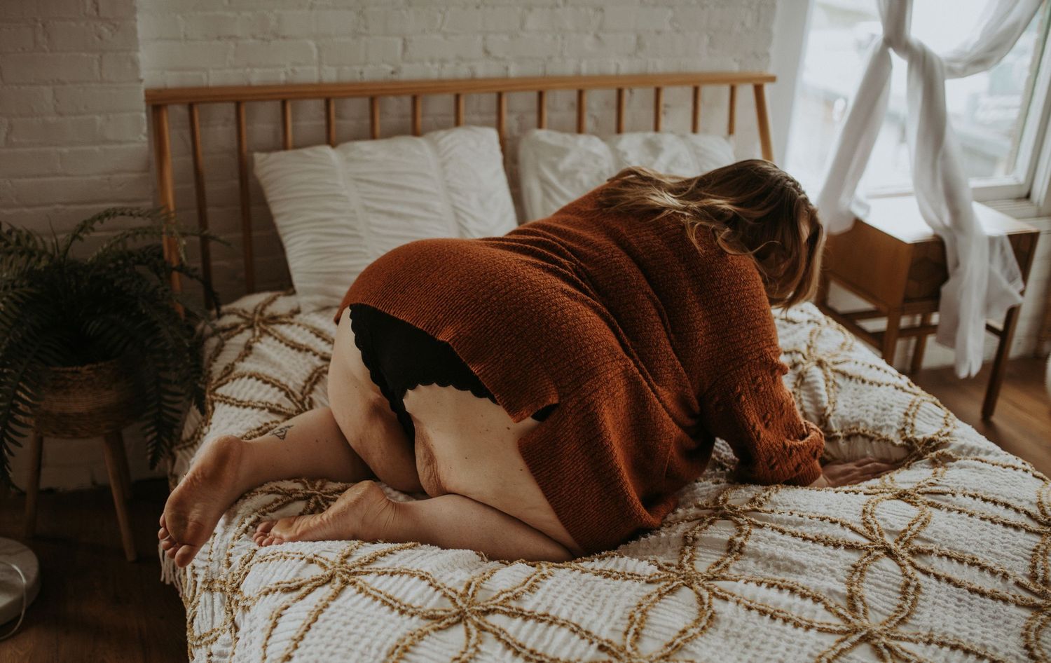A plus-sized woman wearing a rust-colored sweater poses seductively on a bed with patterned duvet in a cozy bedroom setting.