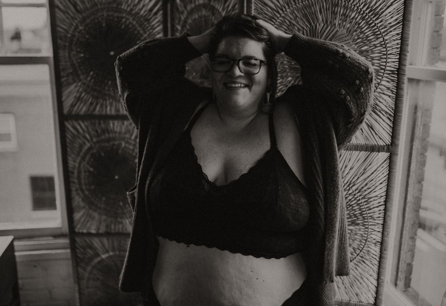 A black and white photo showing a woman in lingerie smiling against a decorative patterned wall divider.