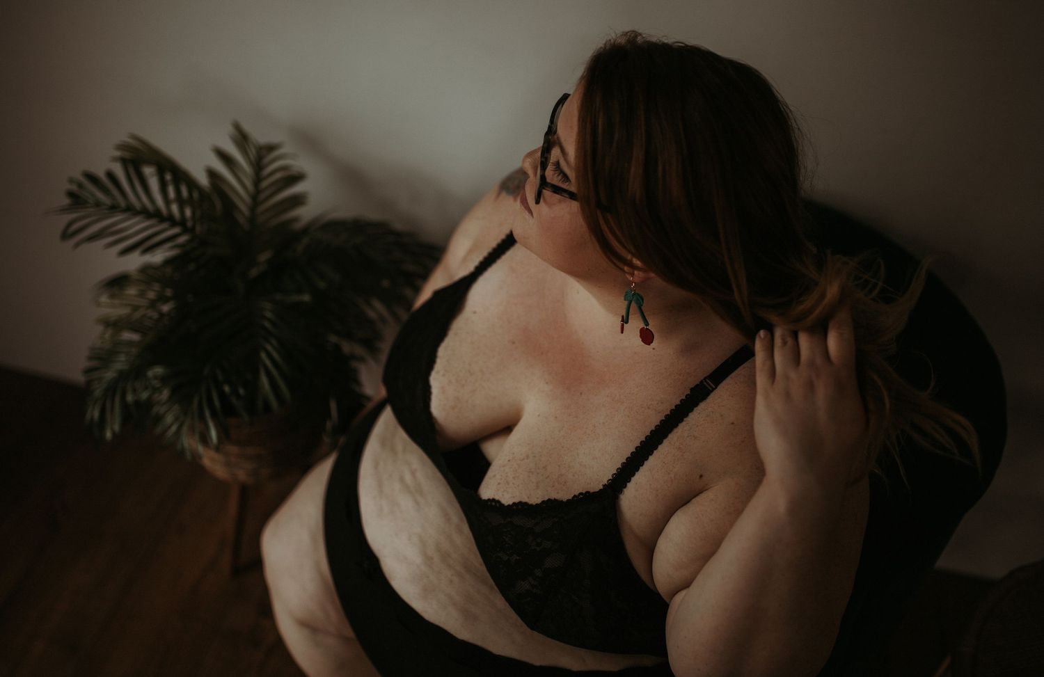 Moody boudoir photo featuring a woman in black lingerie running her hands through her hair with a houseplant in the background.