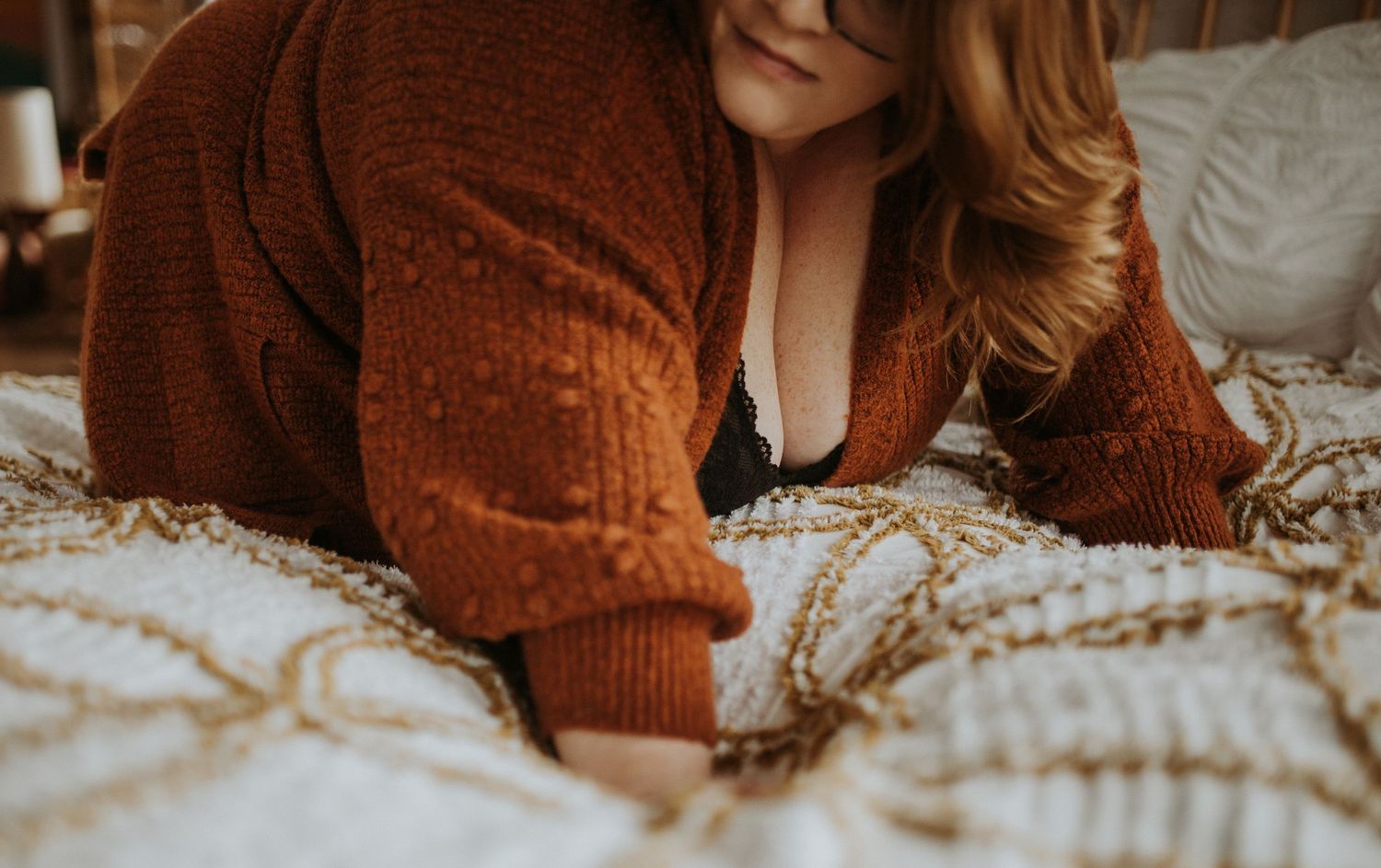 A woman wearing a rust knitted cardigan lies on patterned duvet in a cozy bedroom setting.