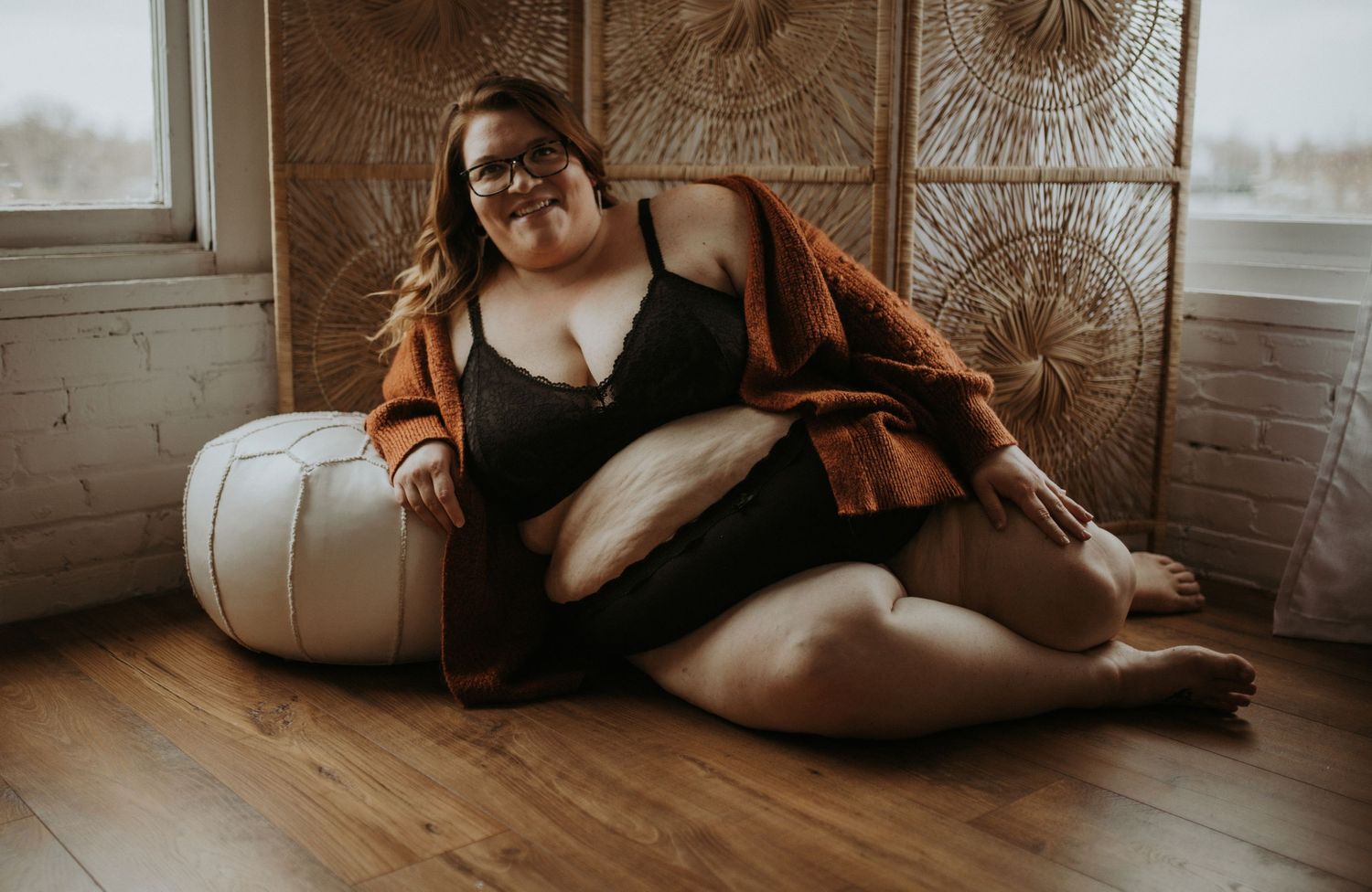 Moody boudoir portrait of person in black dress and brown cardigan reclining on white ottoman near window.