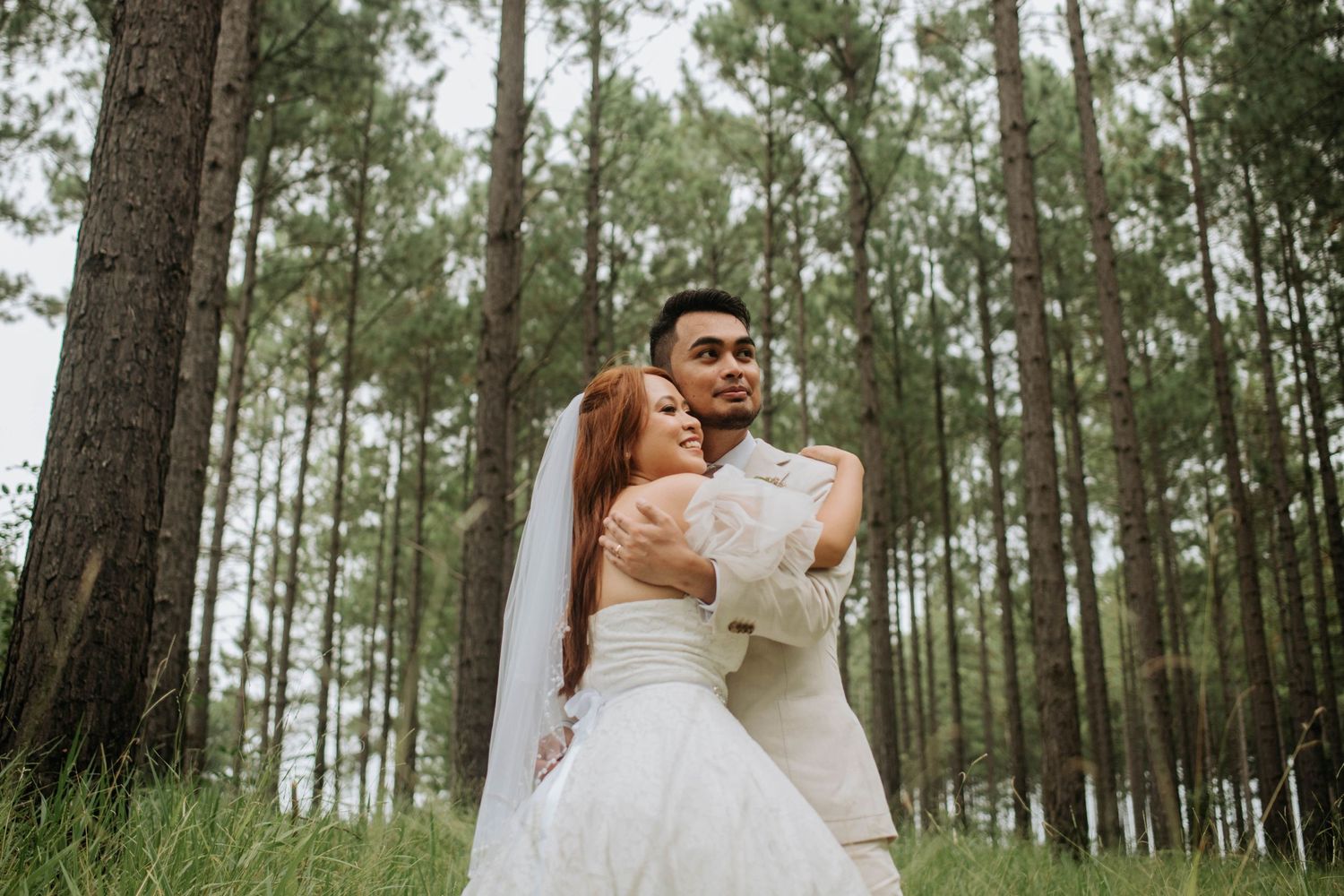 Wedding couple embraces in forest setting surrounded by tall pine trees.