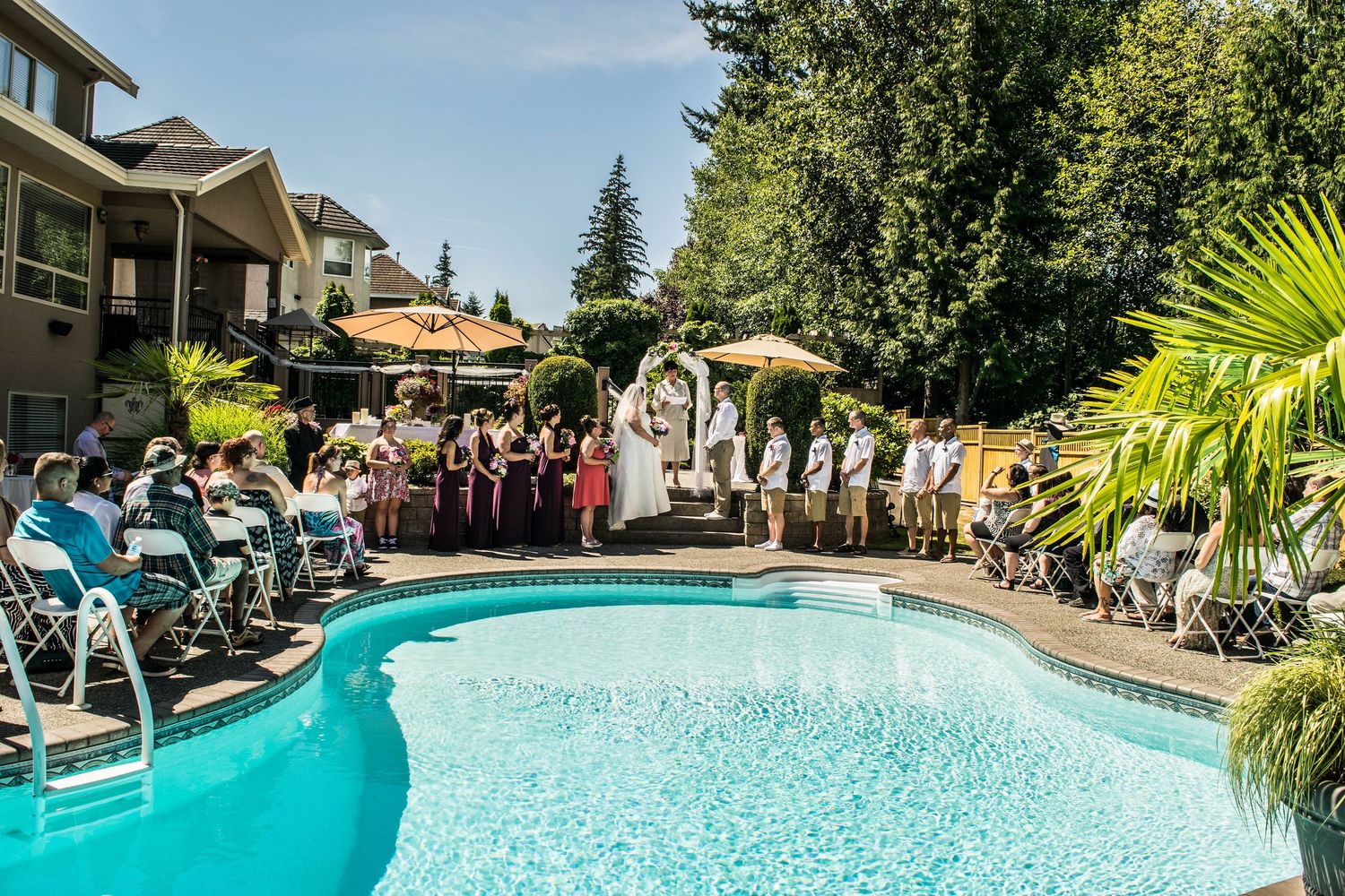 Outdoor wedding ceremony setup around a curved swimming pool with white chairs and tropical palm trees.
