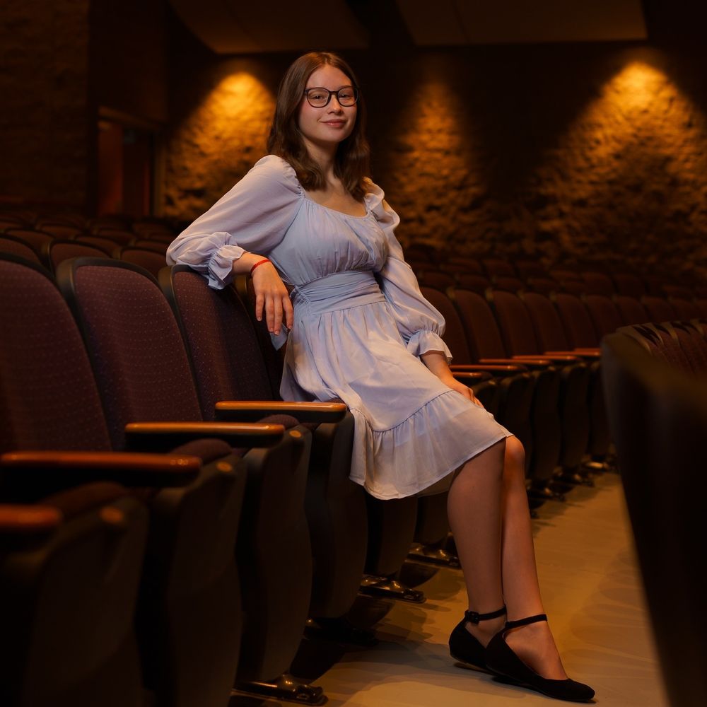 A woman in a light blue dress poses elegantly in theater seating.