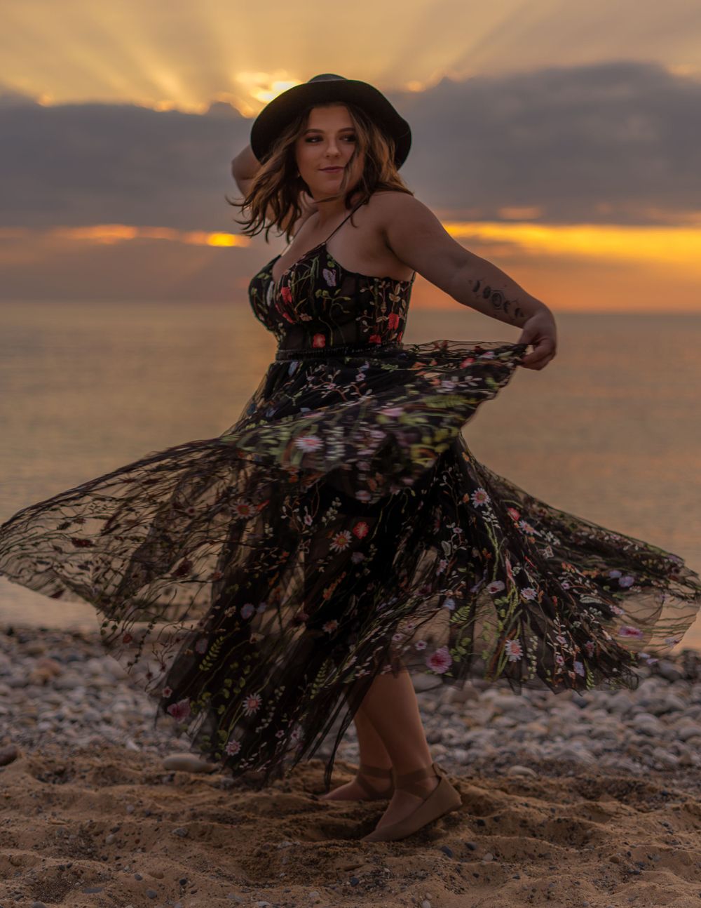 Woman in floral dress and hat twirls on beach during golden sunset.