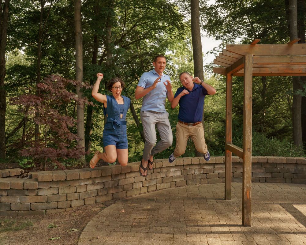 Three people jumping with joy on stone retaining wall in backyard.