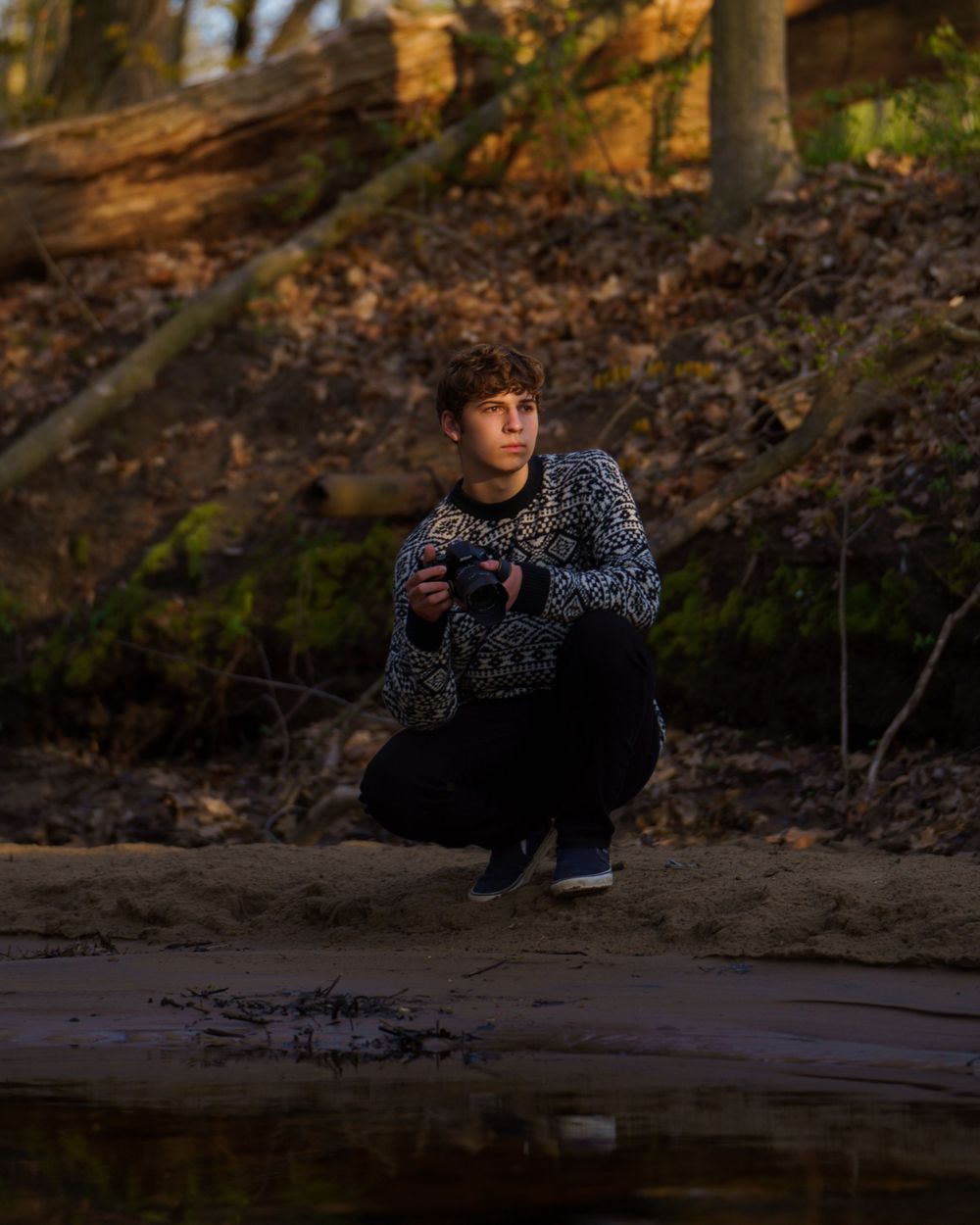 Photographer in patterned sweater crouching by reflective water at sunset in autumn forest setting.