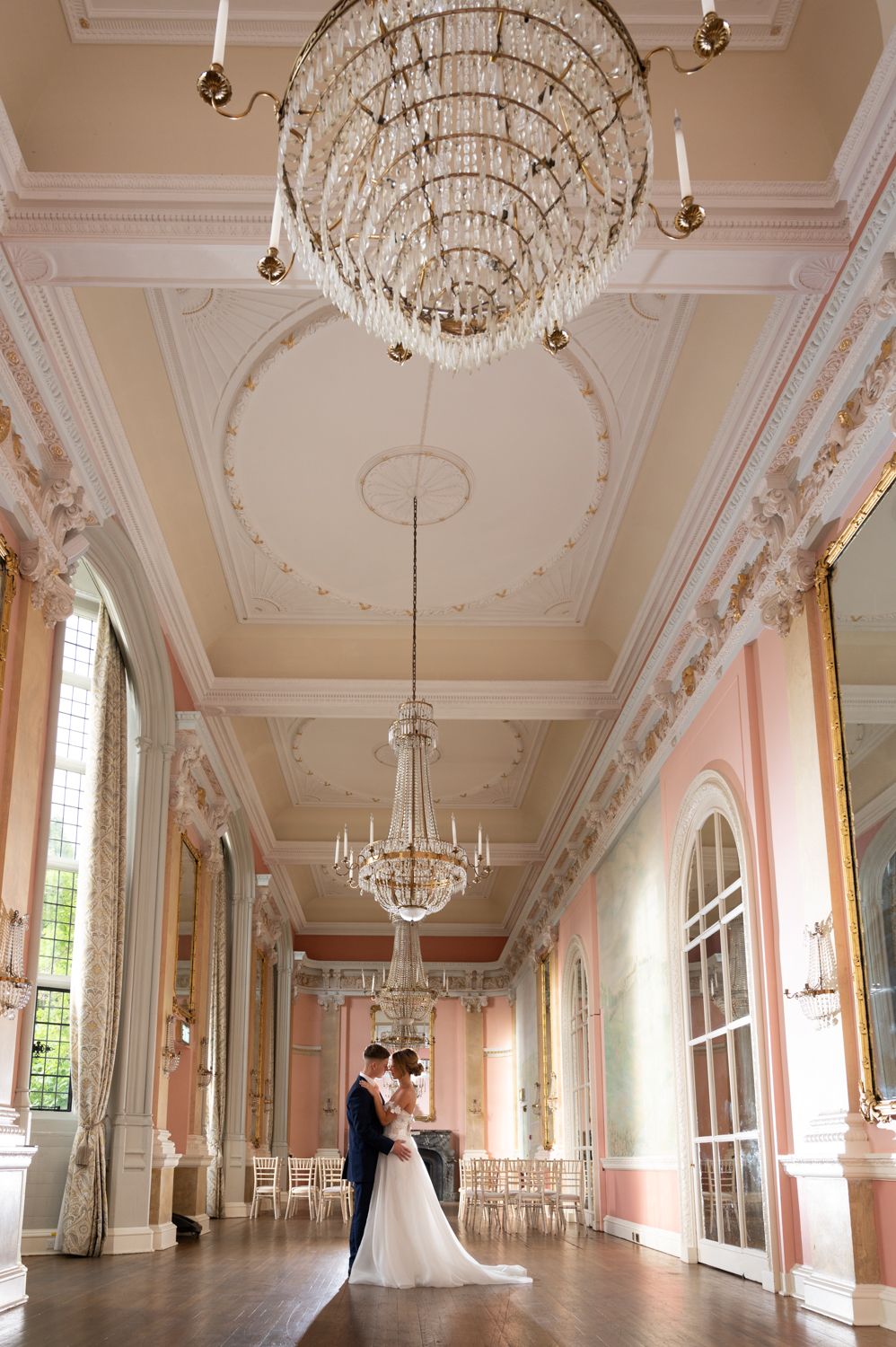 Wedding couple dancing in elegant ballroom with ornate crystal chandelier and pink walls.