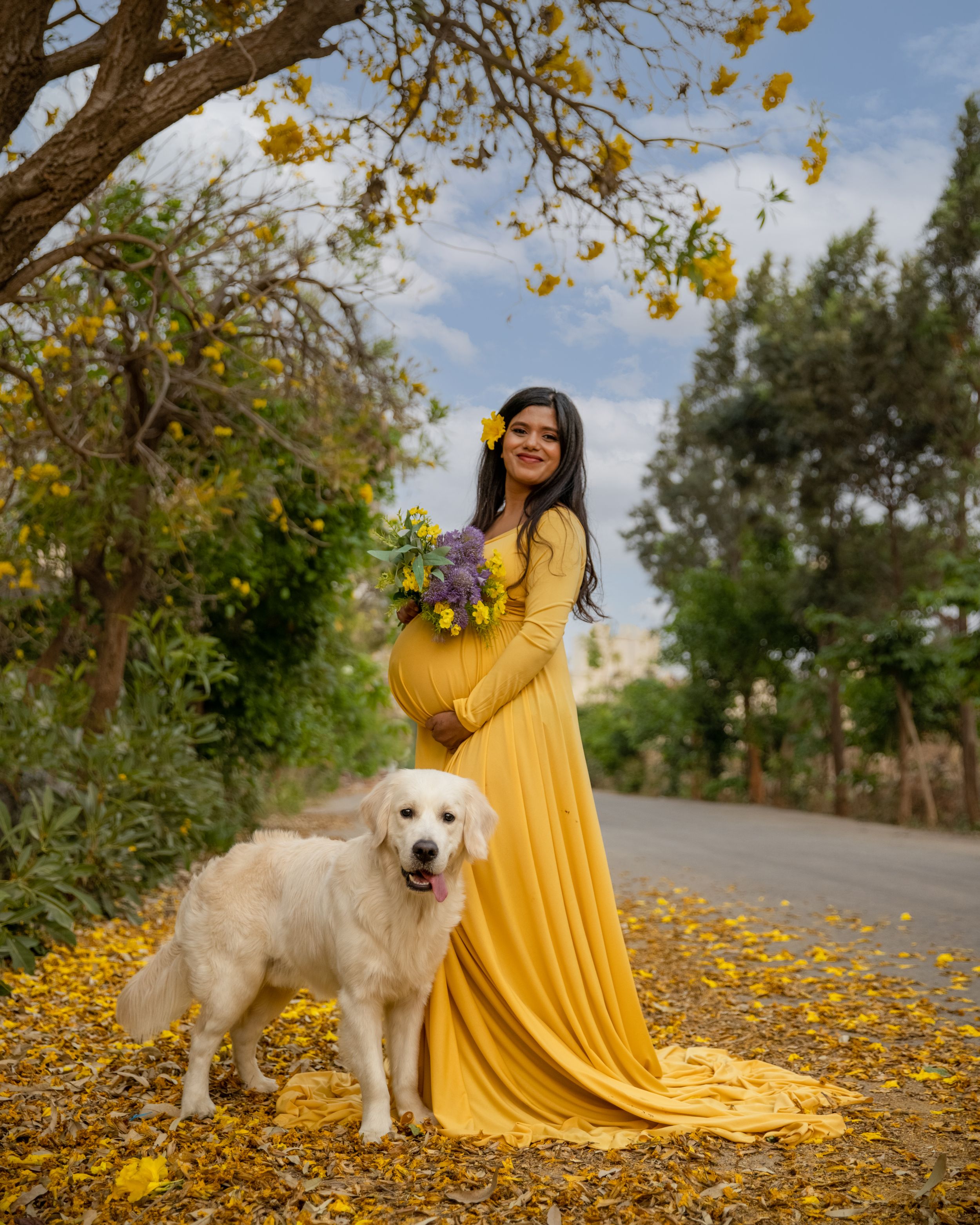 Expectant mother in yellow dress poses outdoors with golden retriever among fall leaves.