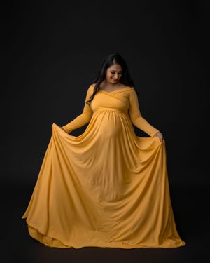 Woman in flowing mustard yellow maternity gown poses elegantly against dark backdrop.