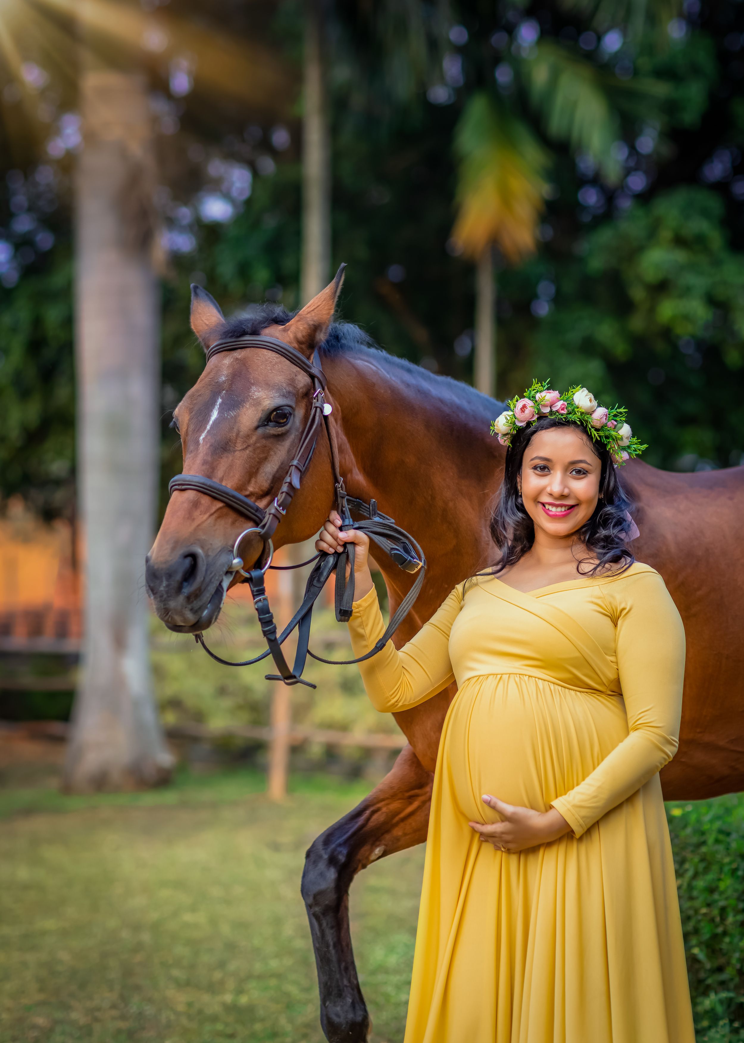 Maternity photo with brown horse featuring yellow dress and floral crown in tropical garden setting.