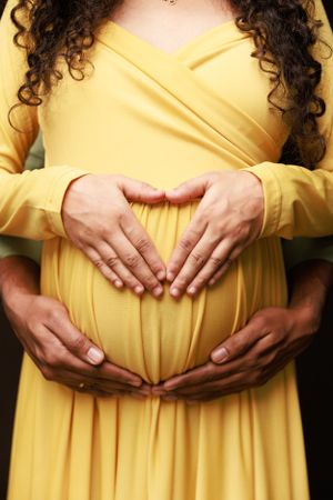 Close-up of hands forming heart shape on pregnant belly in yellow dress.