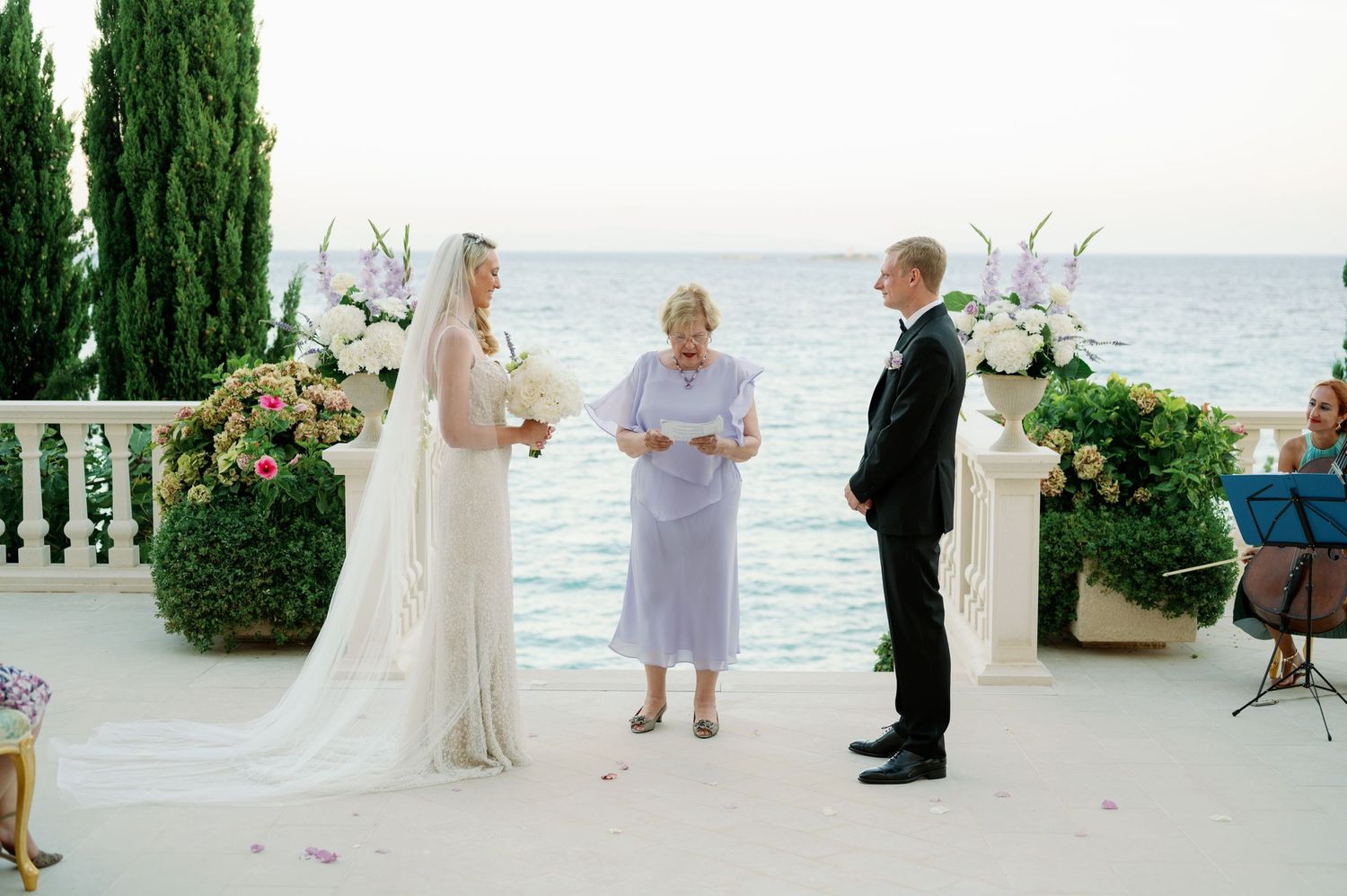 Oceanside wedding ceremony with white floral arrangements and cypress trees.