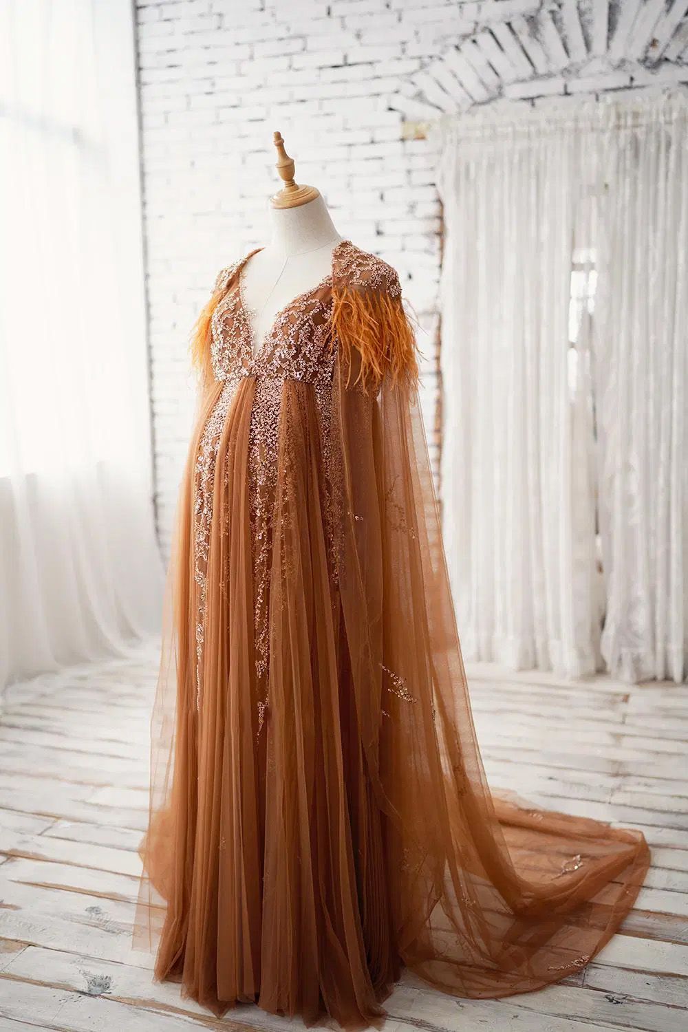 Elegant brown tulle maternity gown with feathers and sequins displayed on mannequin in white studio setting.