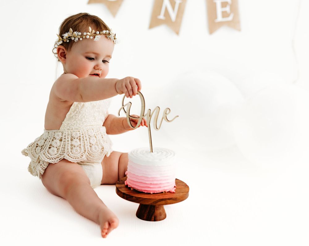 Baby in lace outfit reaches for cake topper during first birthday photo shoot with pink ombre cake.