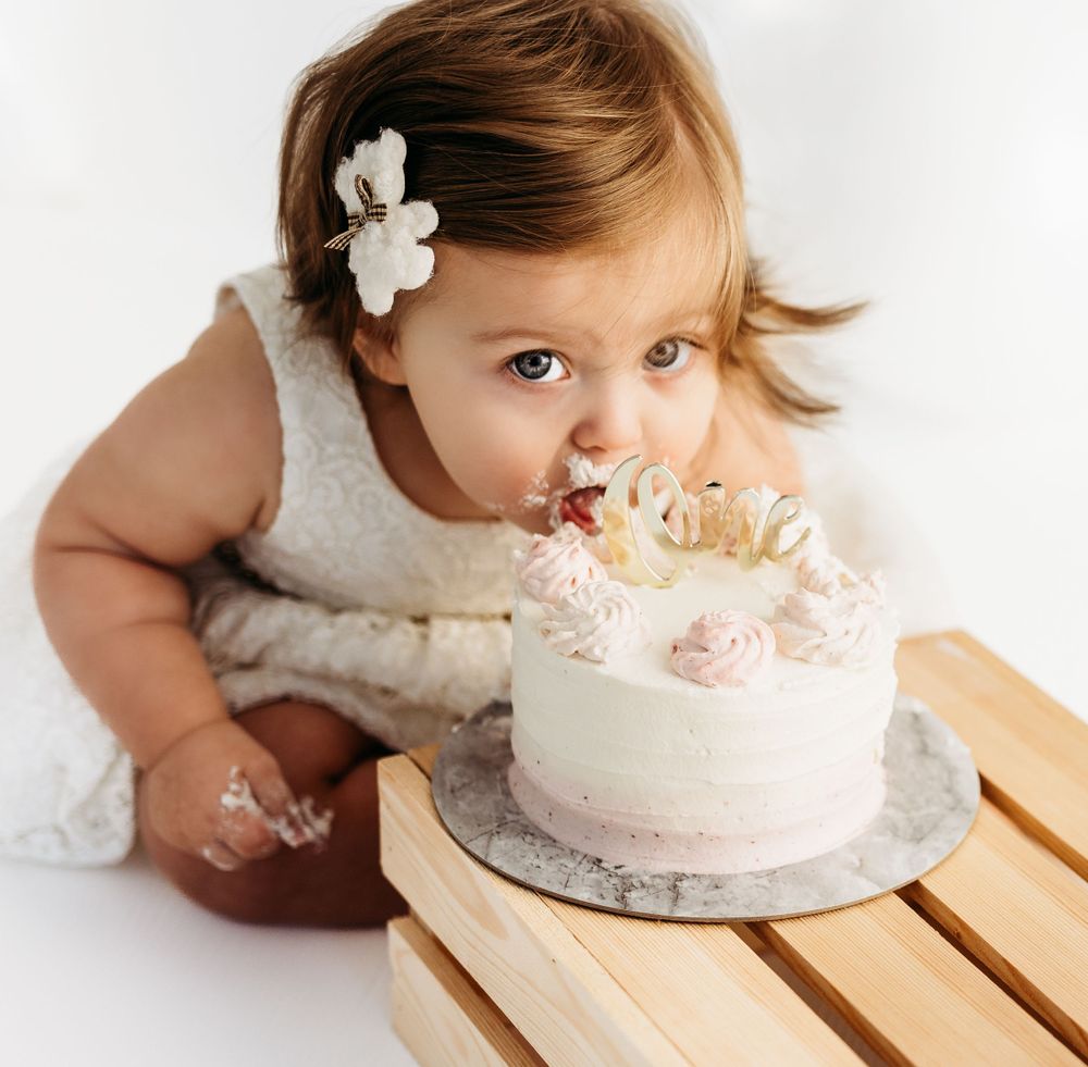 Baby in white dress with hair bow leaning over to taste a decorated pink birthday cake. during a session in Fairfax, VA.