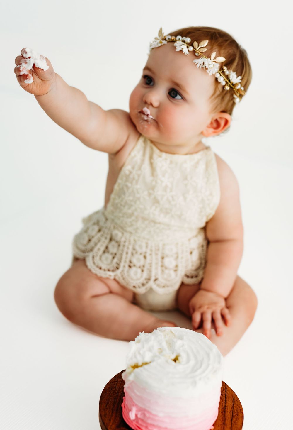 Baby in cream lace dress and floral crown reaches for a pink ombre smash cake. during a session in Fairfax, VA.
